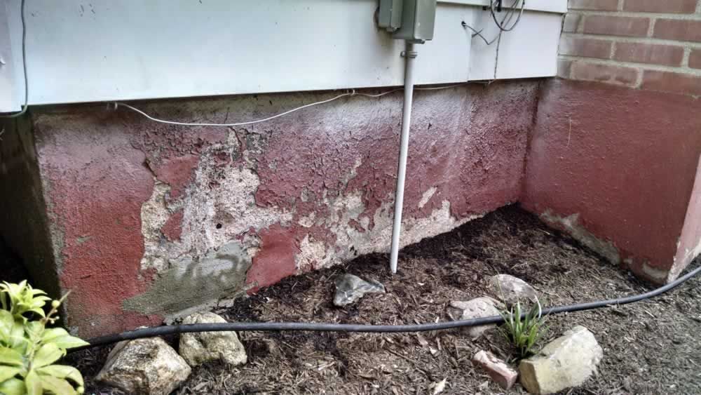 A brick house with a red foundation is sitting next to a hose in the dirt.