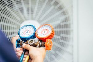 Two hand-held pressure gauges pointed at a large fan, with blue and orange dials in front of a metal grille.