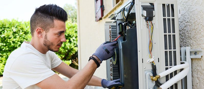Technician inspecting outdoor air conditioning unit with tools, wearing gloves beside a house