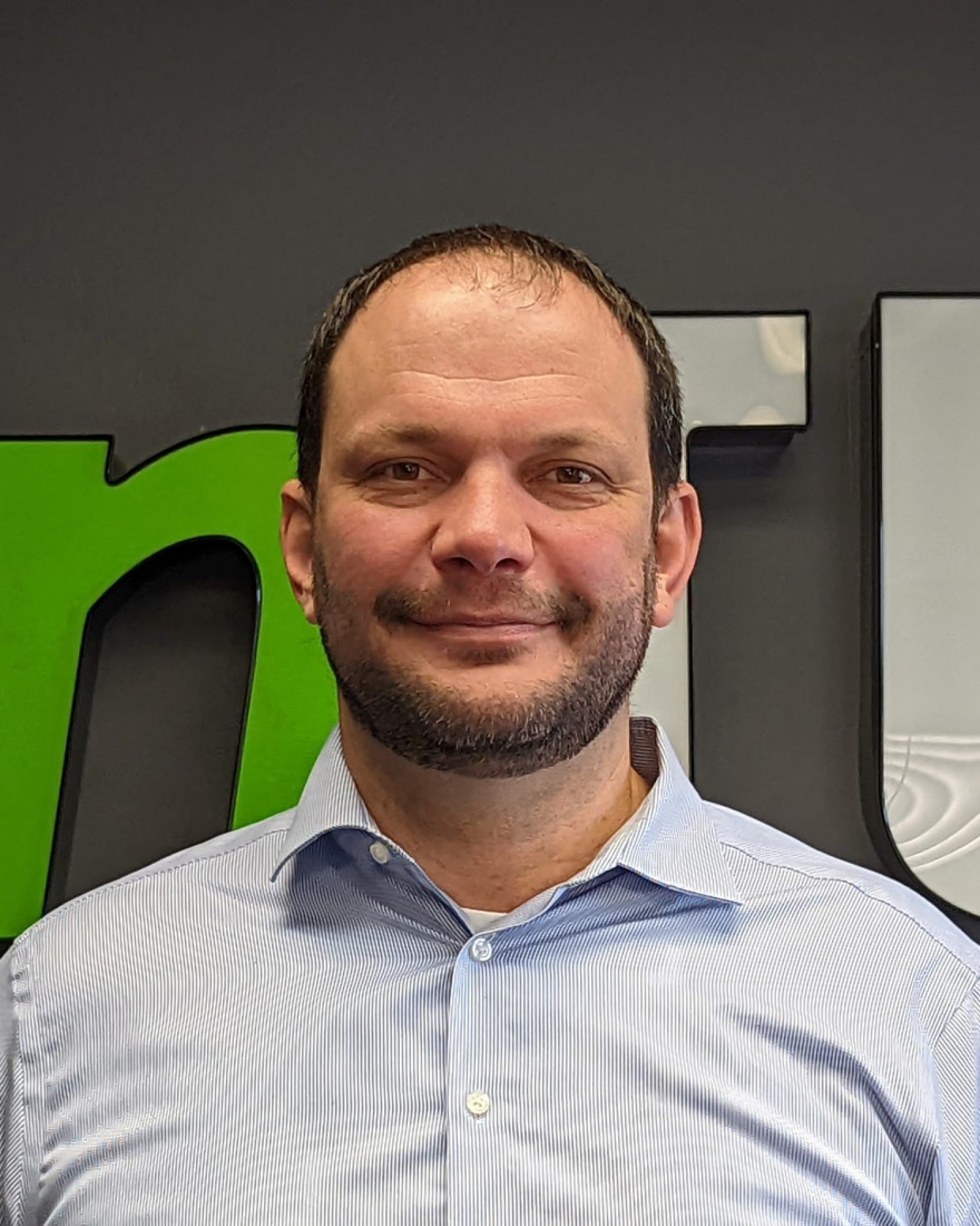 Smiling man in a blue striped shirt standing indoors against a dark wall with green and white signage