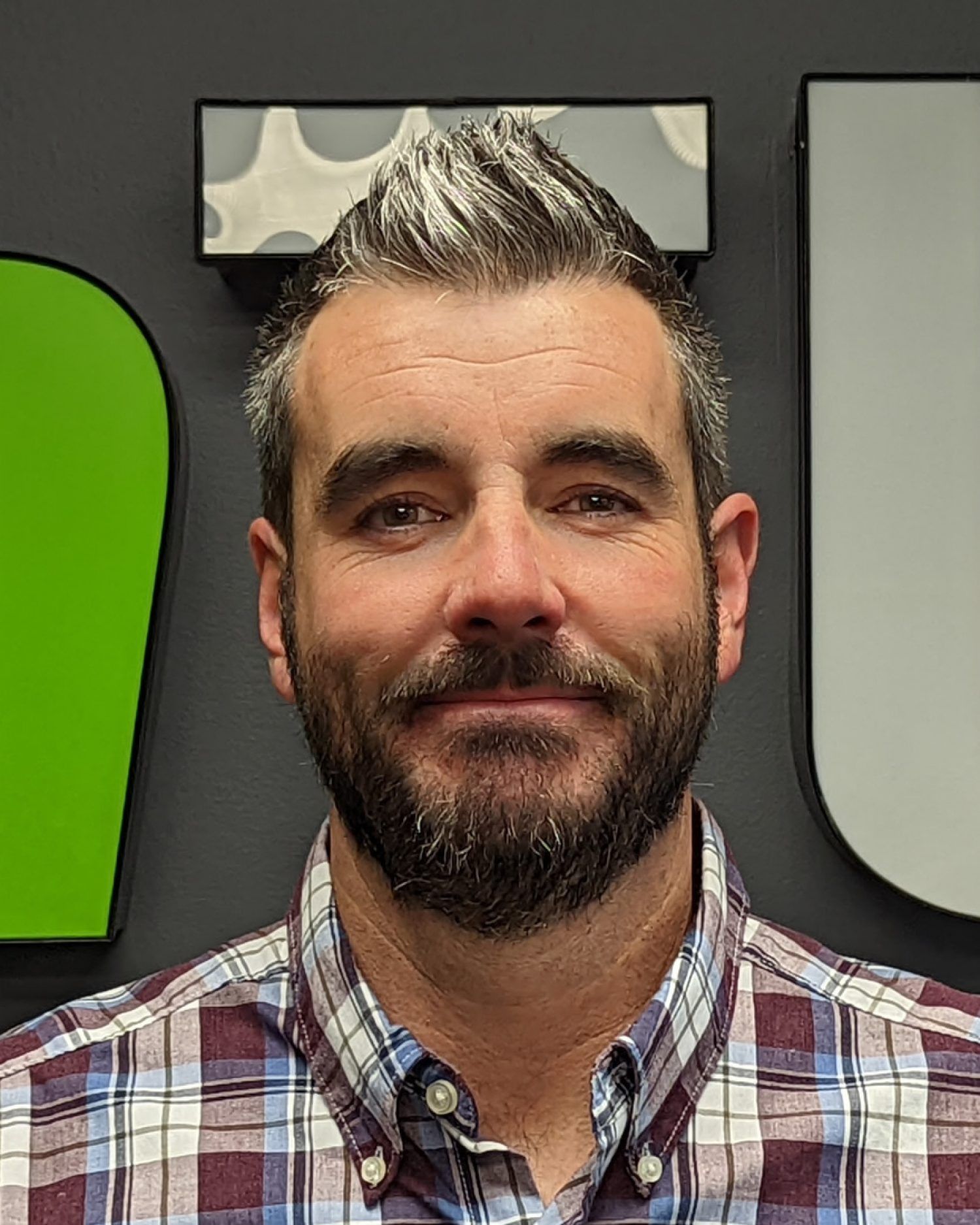 Smiling man with gray-streaked hair and beard in a plaid shirt against a gray wall with green and white panels