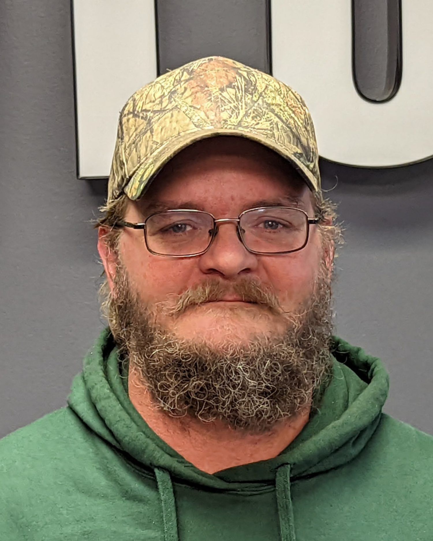 Man in green hoodie and camouflage cap, wearing glasses and a beard, standing against a gray wall.