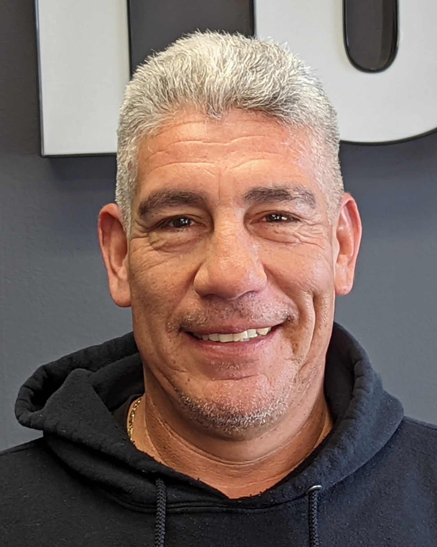 Smiling person in a black hoodie against a gray wall with large white lettering.