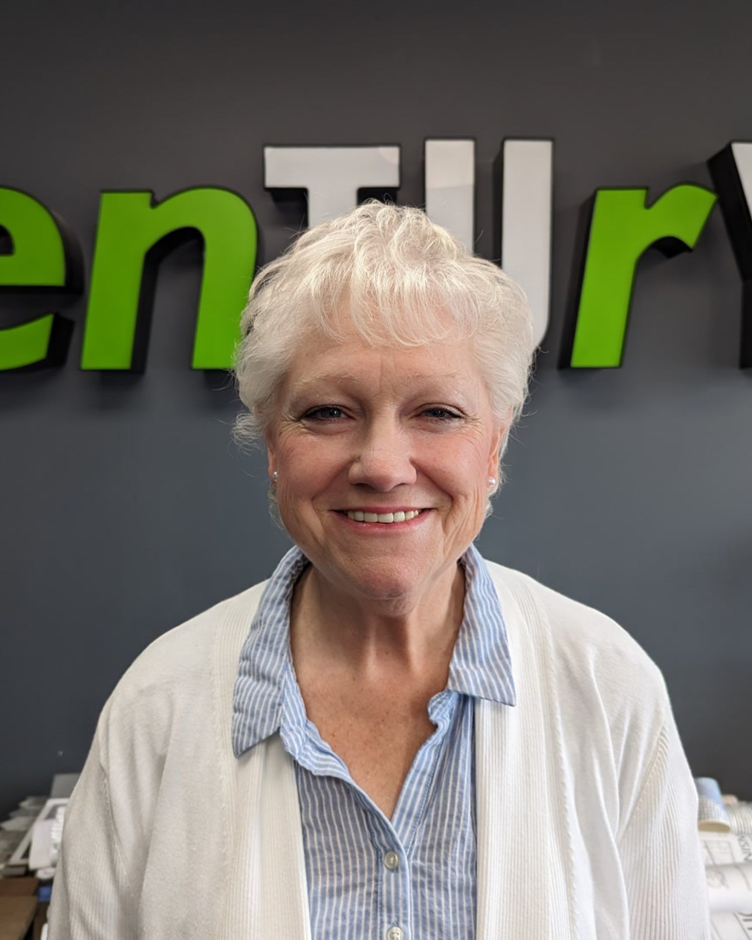 Smiling person with short white hair standing before a wall sign in an office