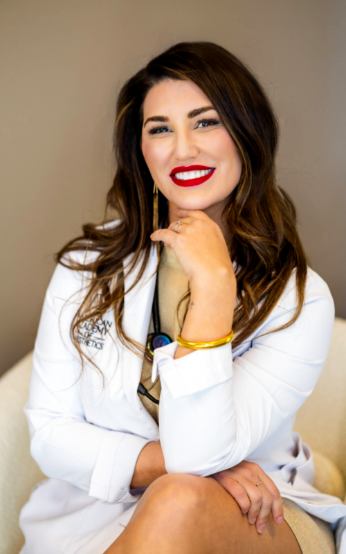 Woman in white outfit smiling, seated with hand on chin against a beige background.