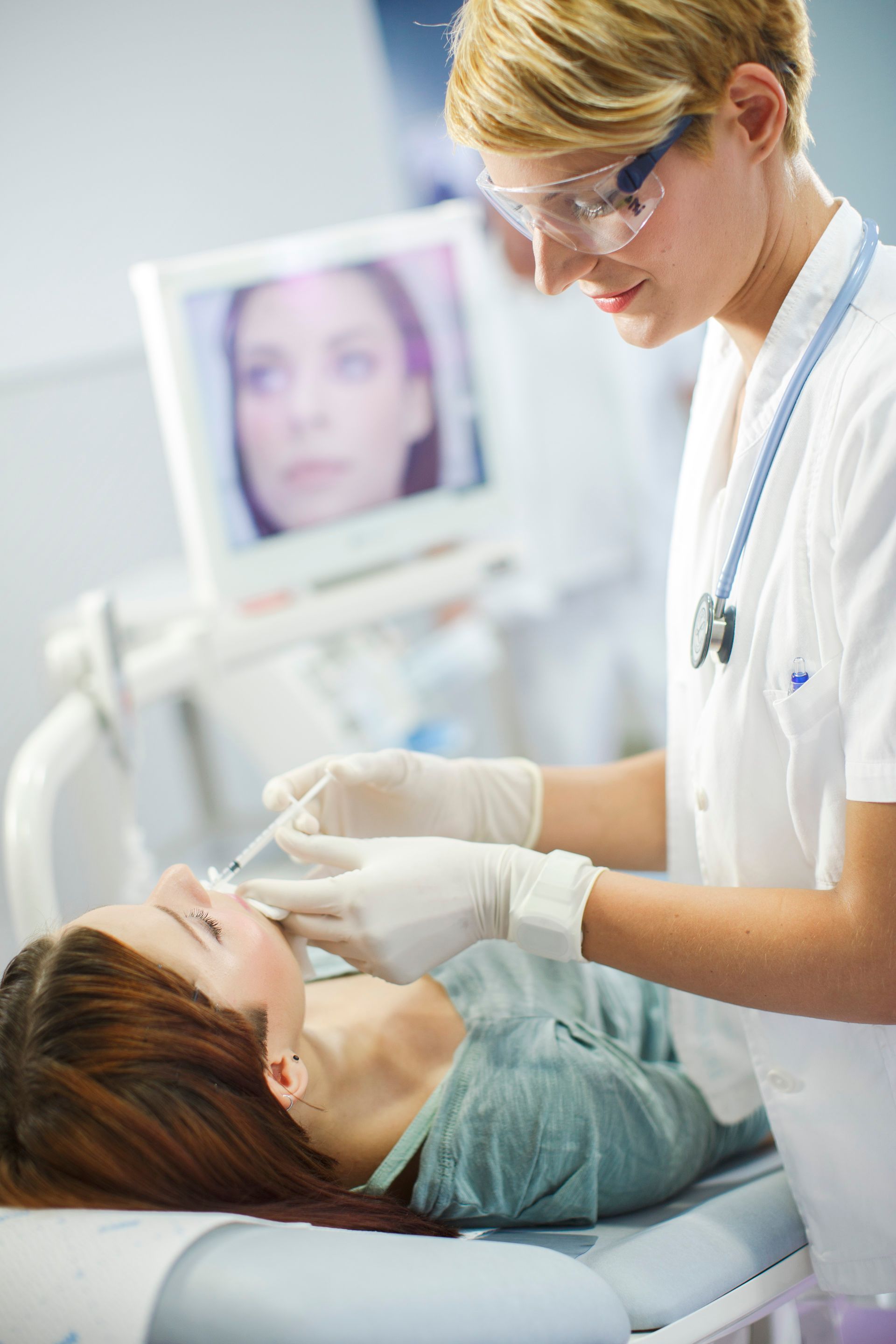 Dentist wearing gloves examines a patient in a dental chair while a monitor glows in the background