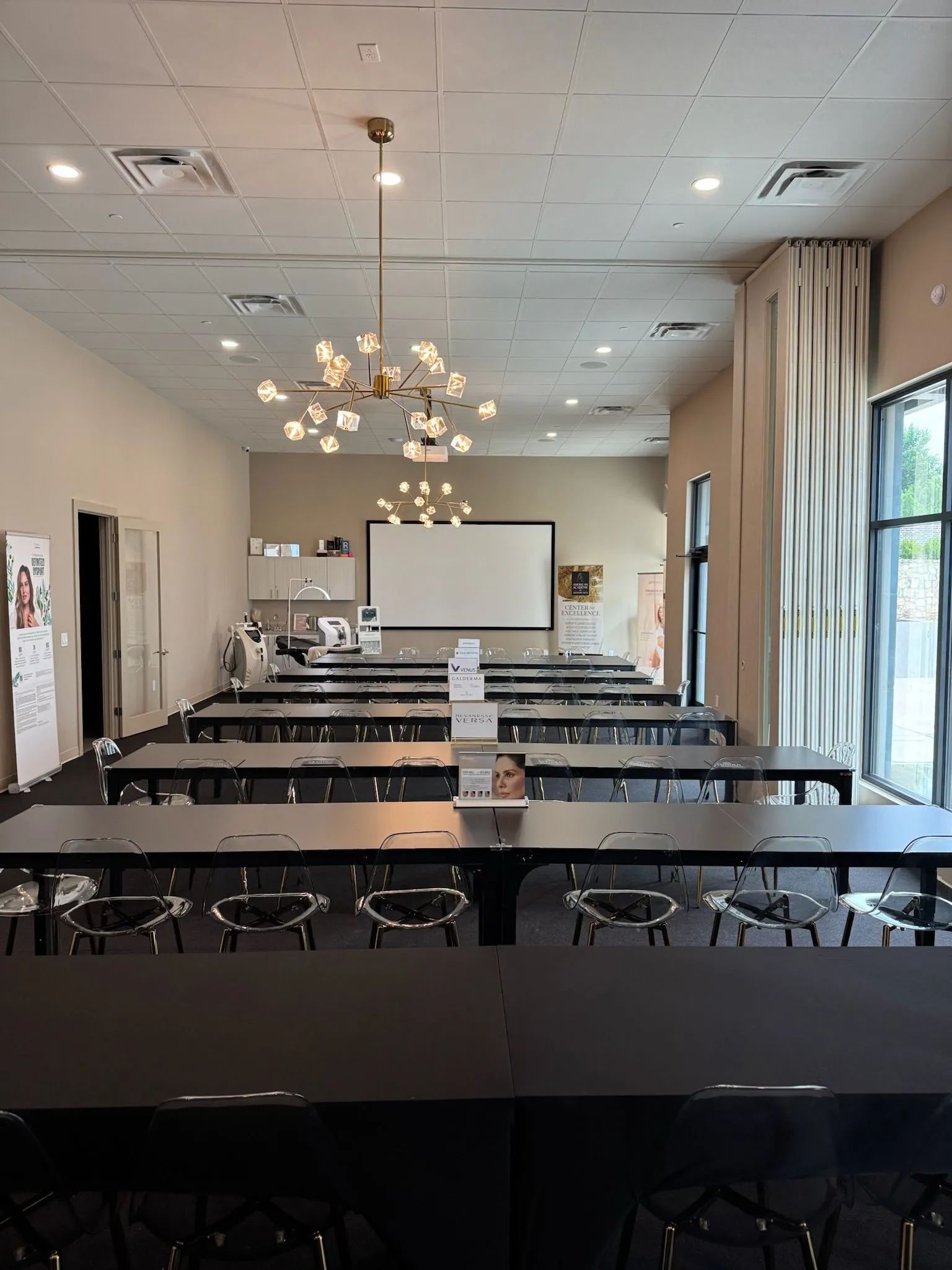 Empty conference room with rows of tables and chairs, projector screen, chandelier, and large windows.