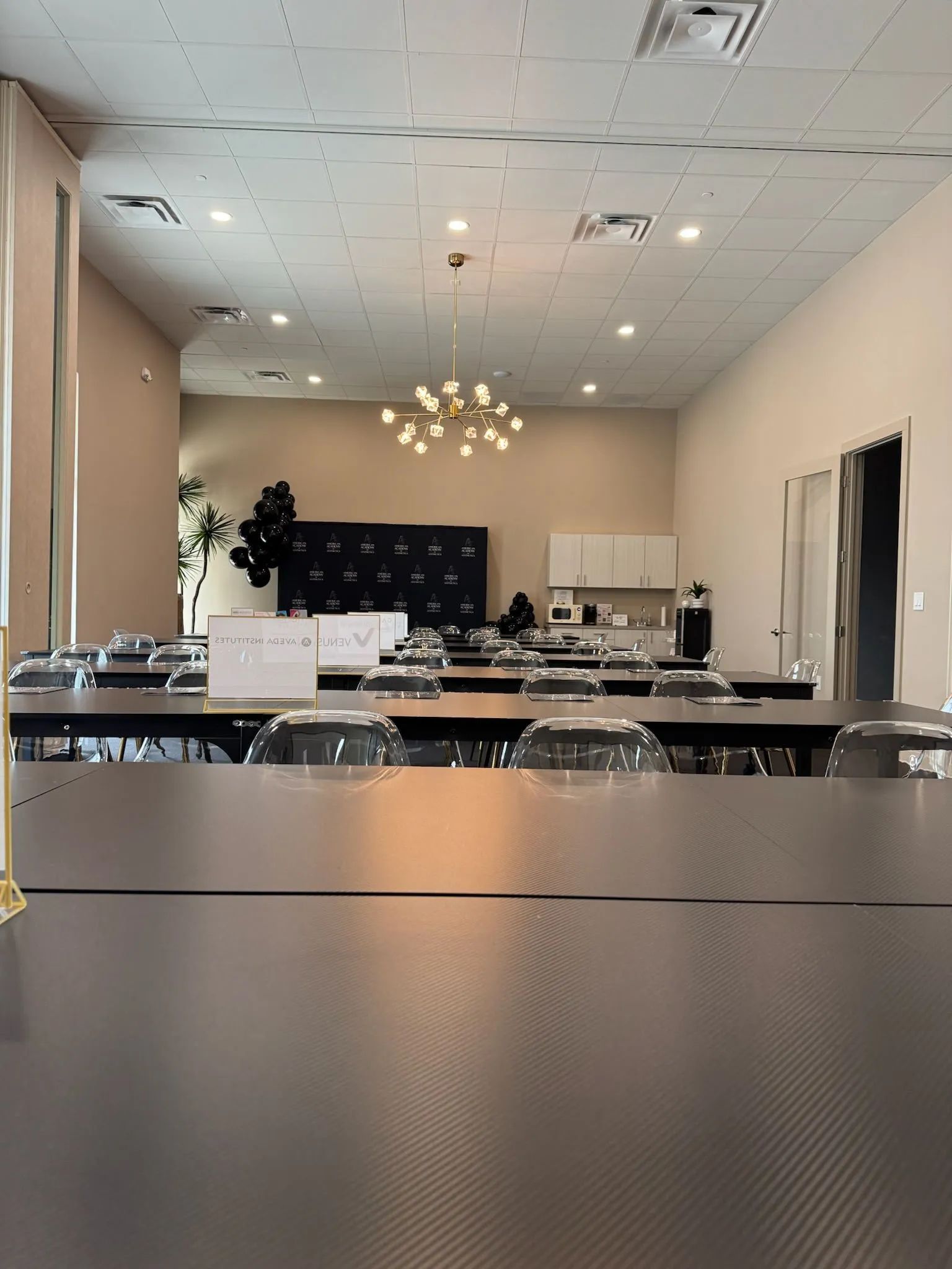 Empty modern event room with tables and chairs under a chandelier, beige walls and ceiling tiles.