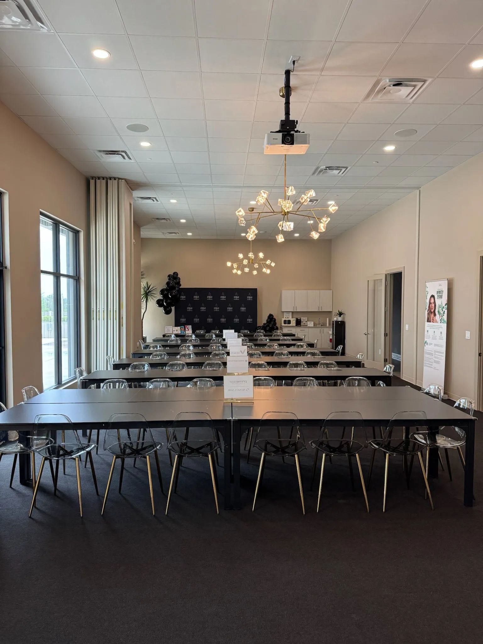 Conference room with long tables, black chairs, chandeliers, and large windows along the left wall