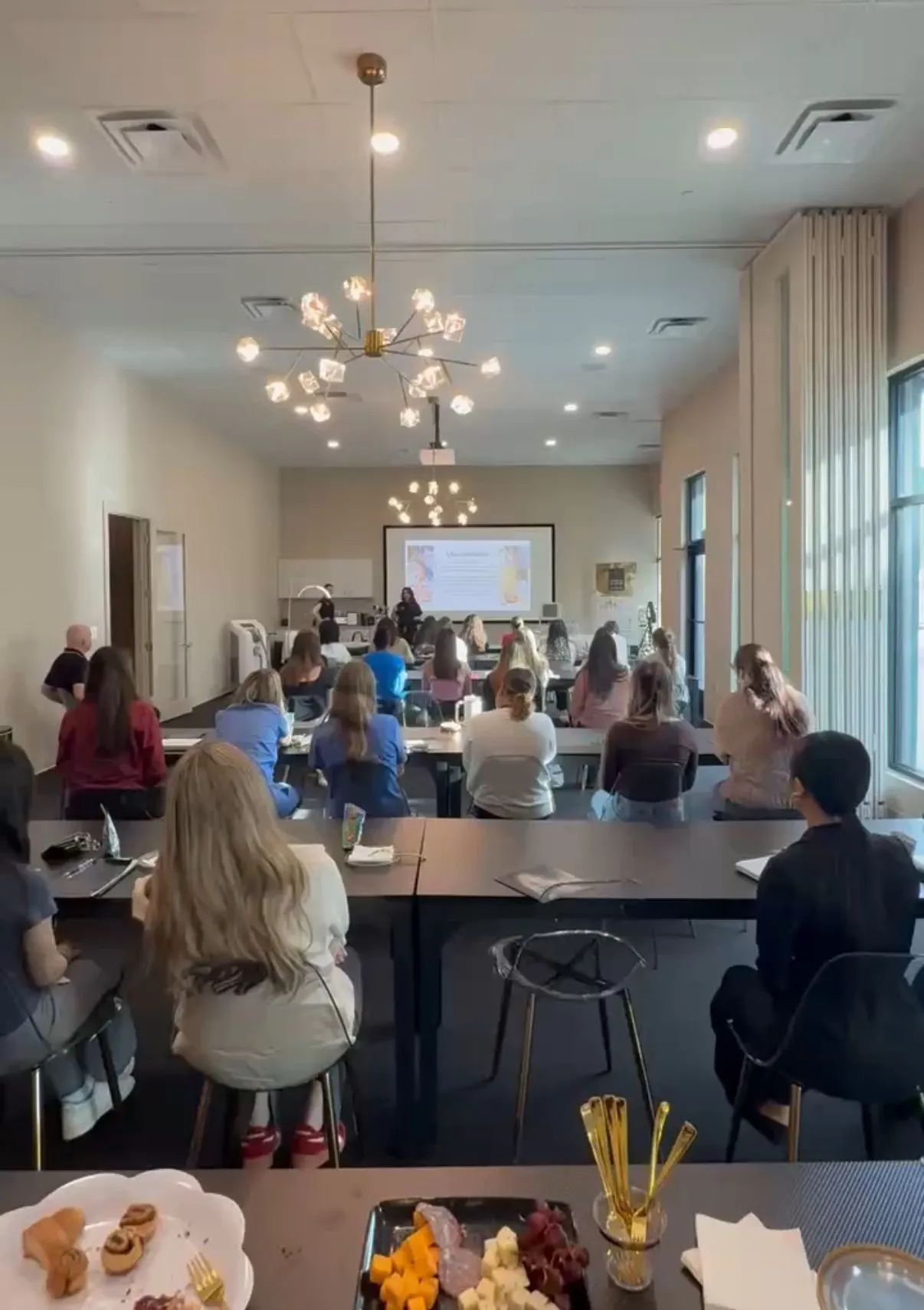 Classroom workshop with people seated at tables facing a projected presentation in a bright room