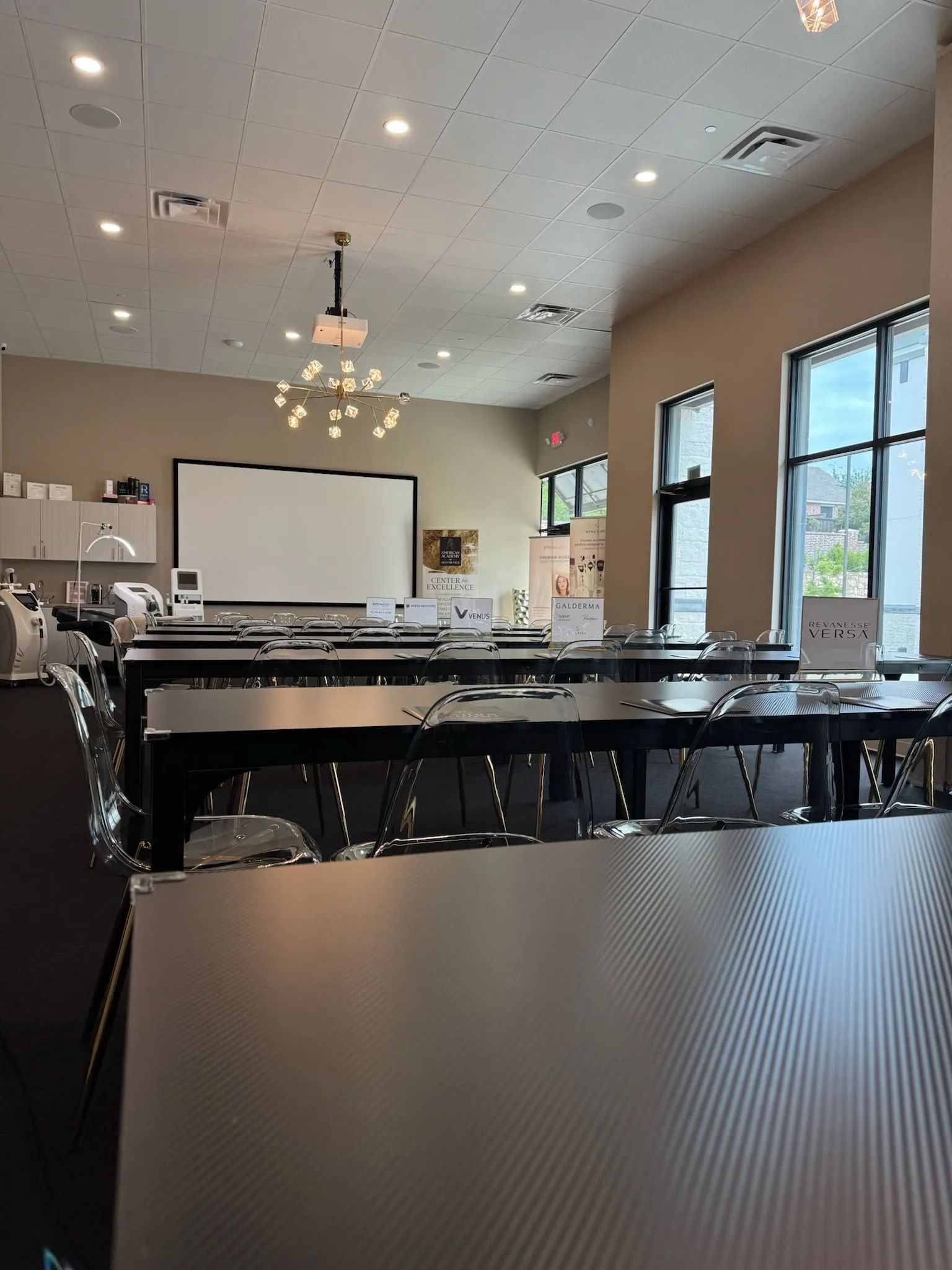 Empty banquet hall with tables, projector screen, chandeliers, and large windows.