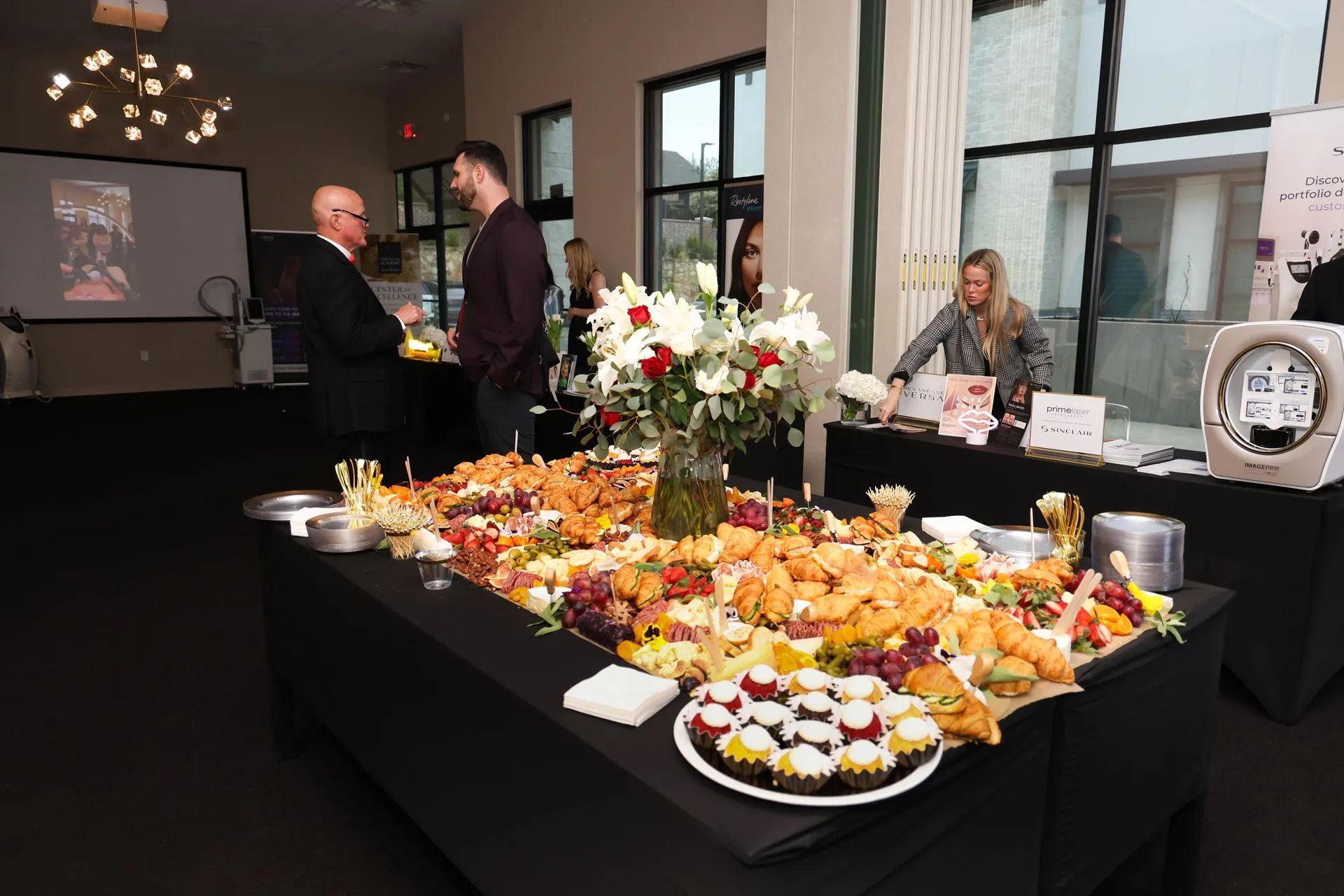 Catered event buffet table with pastries, fruit, flowers, and people mingling in a modern room
