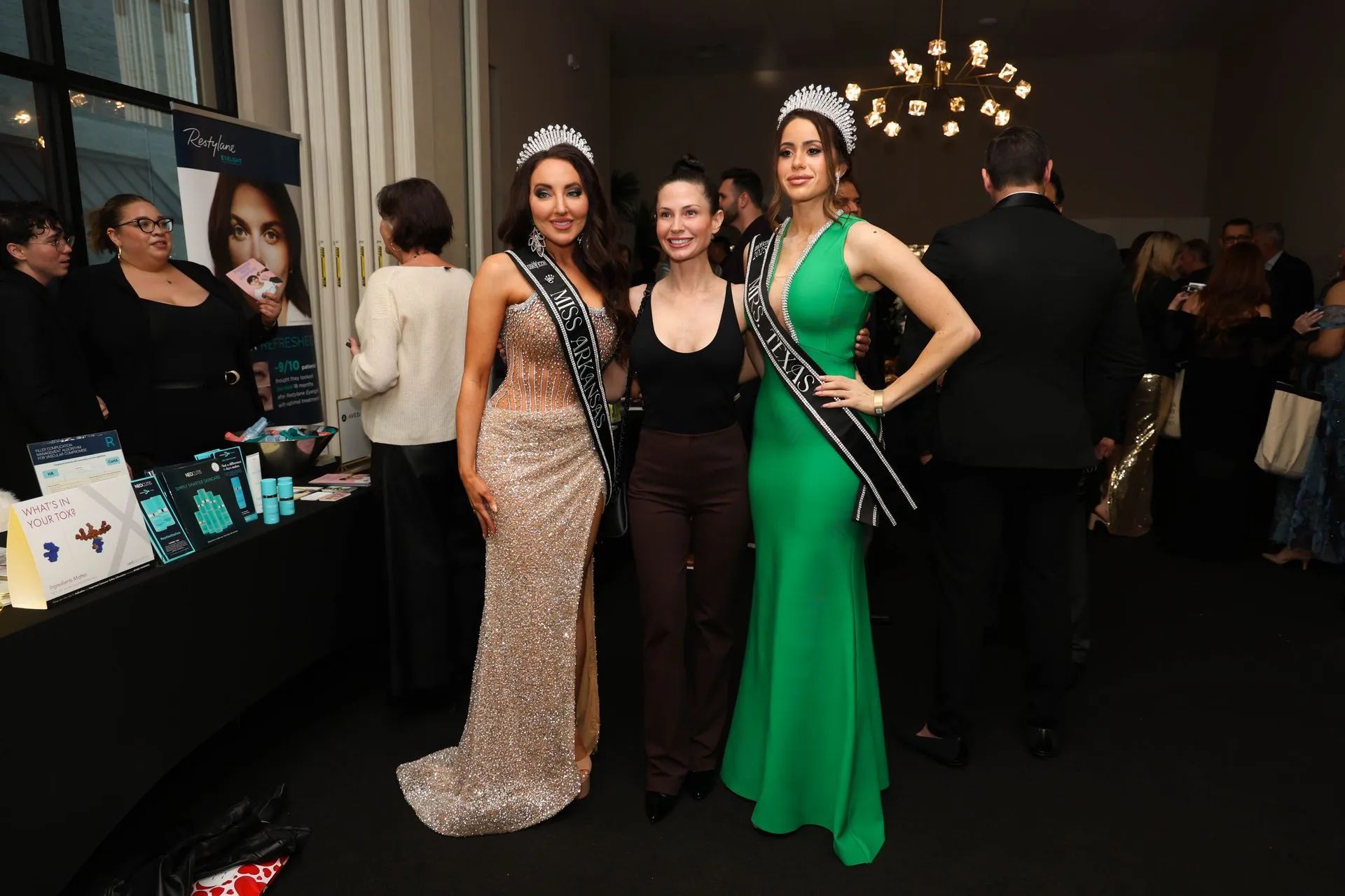 Three women posing at a formal event, one in a beige gown, one in black, one in green with a sash.