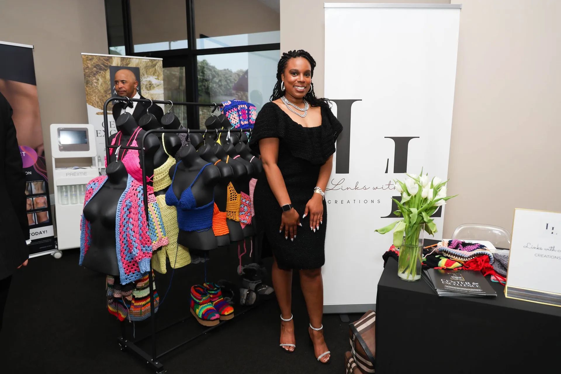 Woman in a black dress standing in an office beside a clothing rack with colorful garments and accessories.