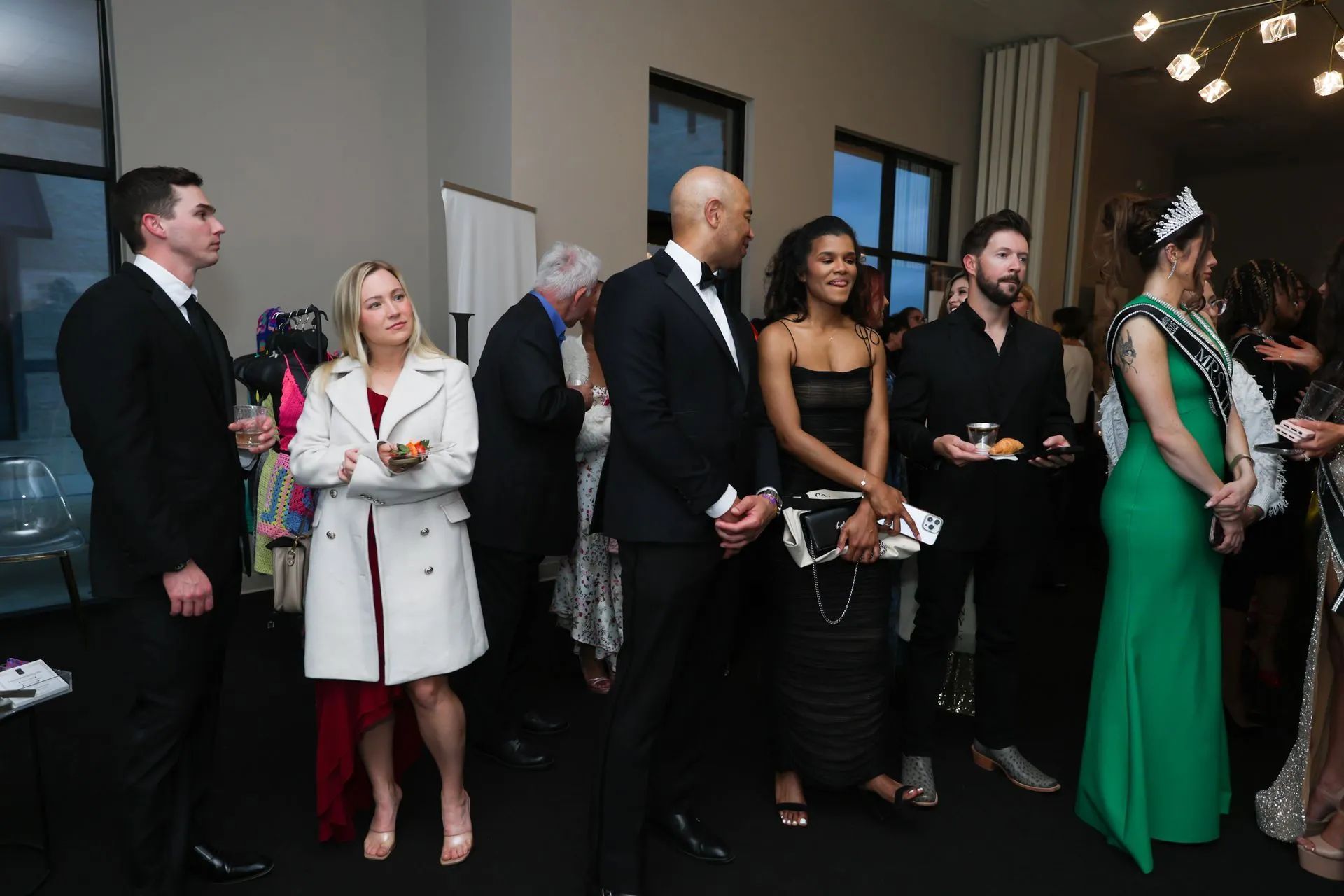 People standing in formal attire at an indoor event, some holding drinks and chatting near a doorway.