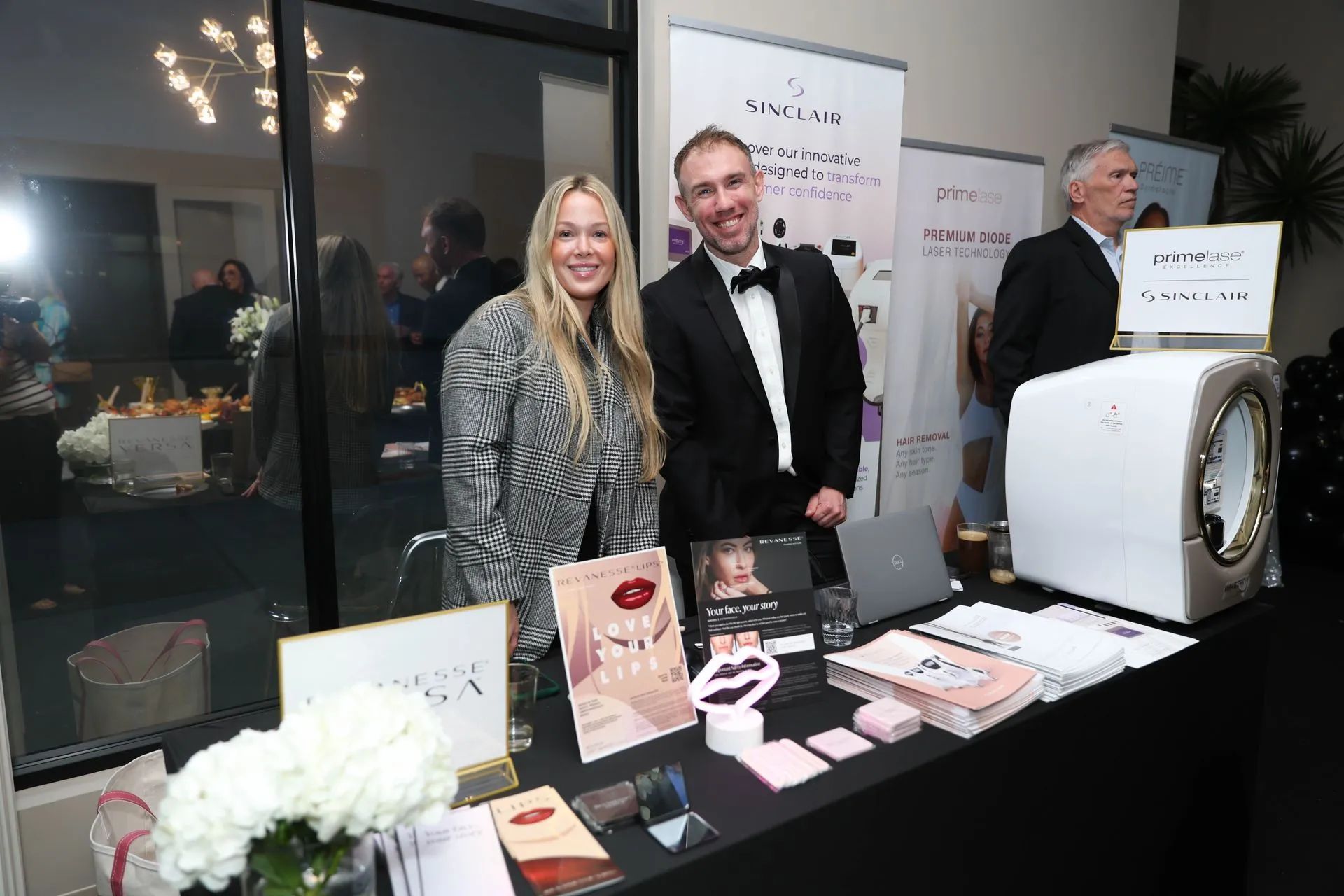 Two people smiling behind a display table at an indoor event booth with products and a large white machine