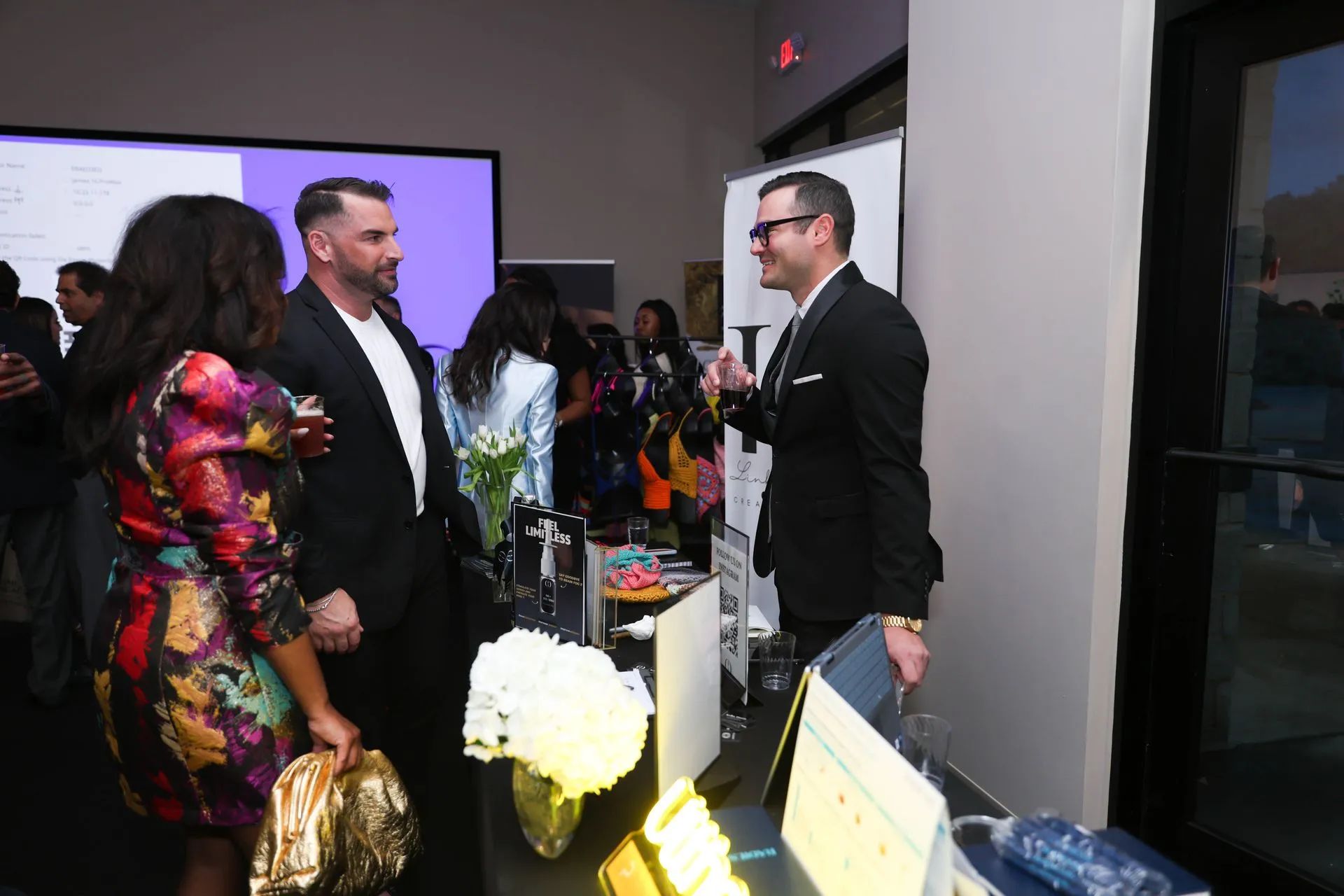 People networking at a reception, with drinks and a presentation screen in the background.