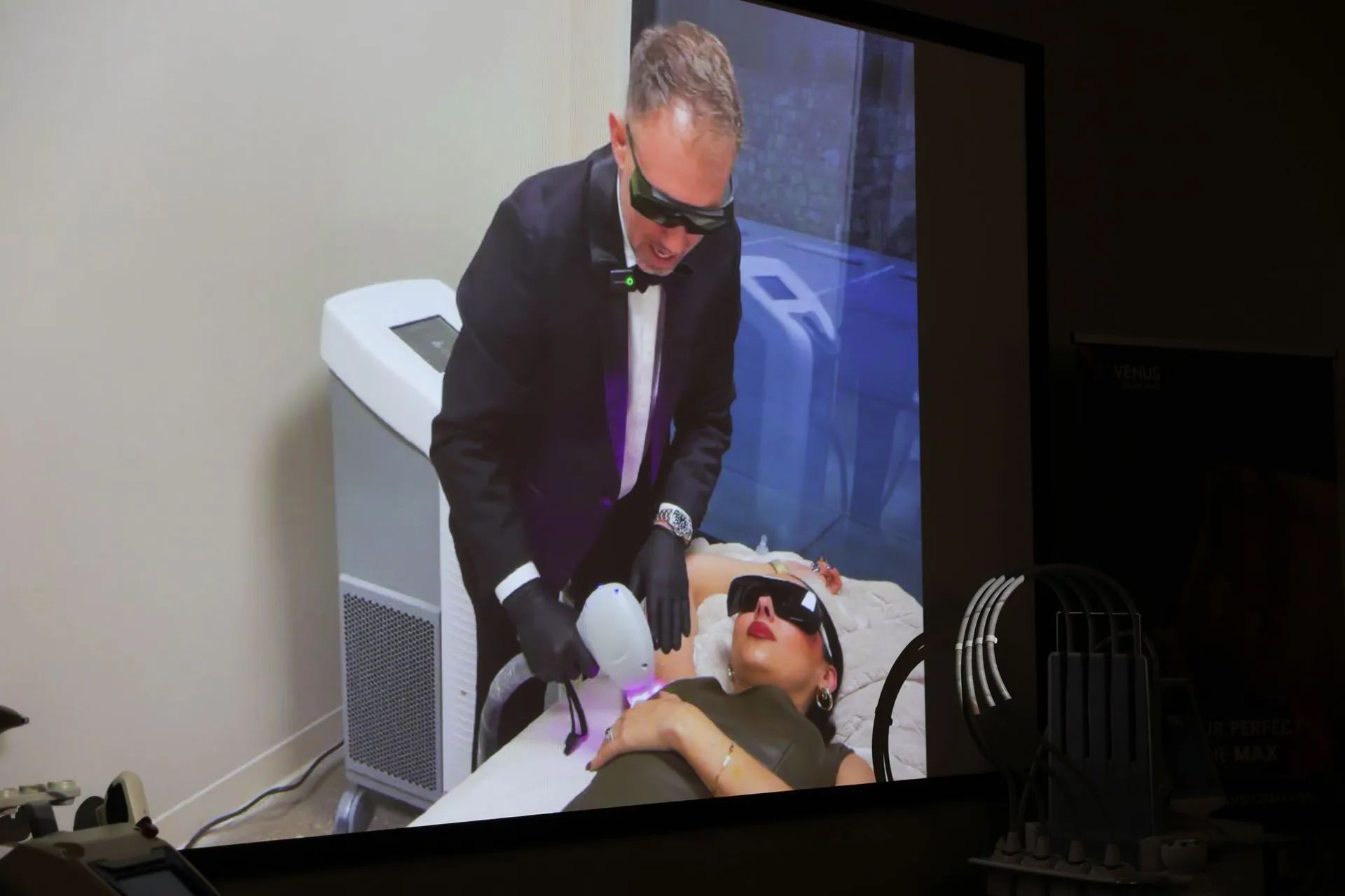 Doctor in dark suit and gloves performing a procedure on a patient lying on a treatment bed under a projected screen