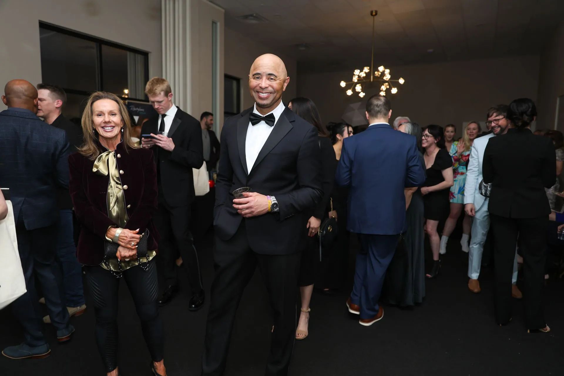 People socializing in a formal indoor reception, with a man in a tuxedo holding a drink in the foreground.