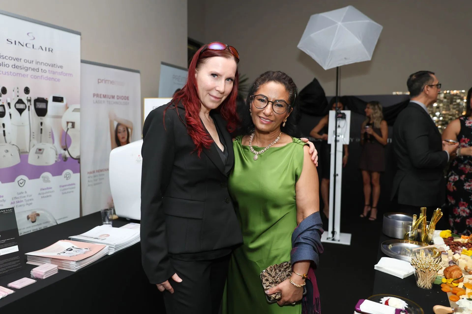 Two women posing together at an indoor event booth, one in black and one in green