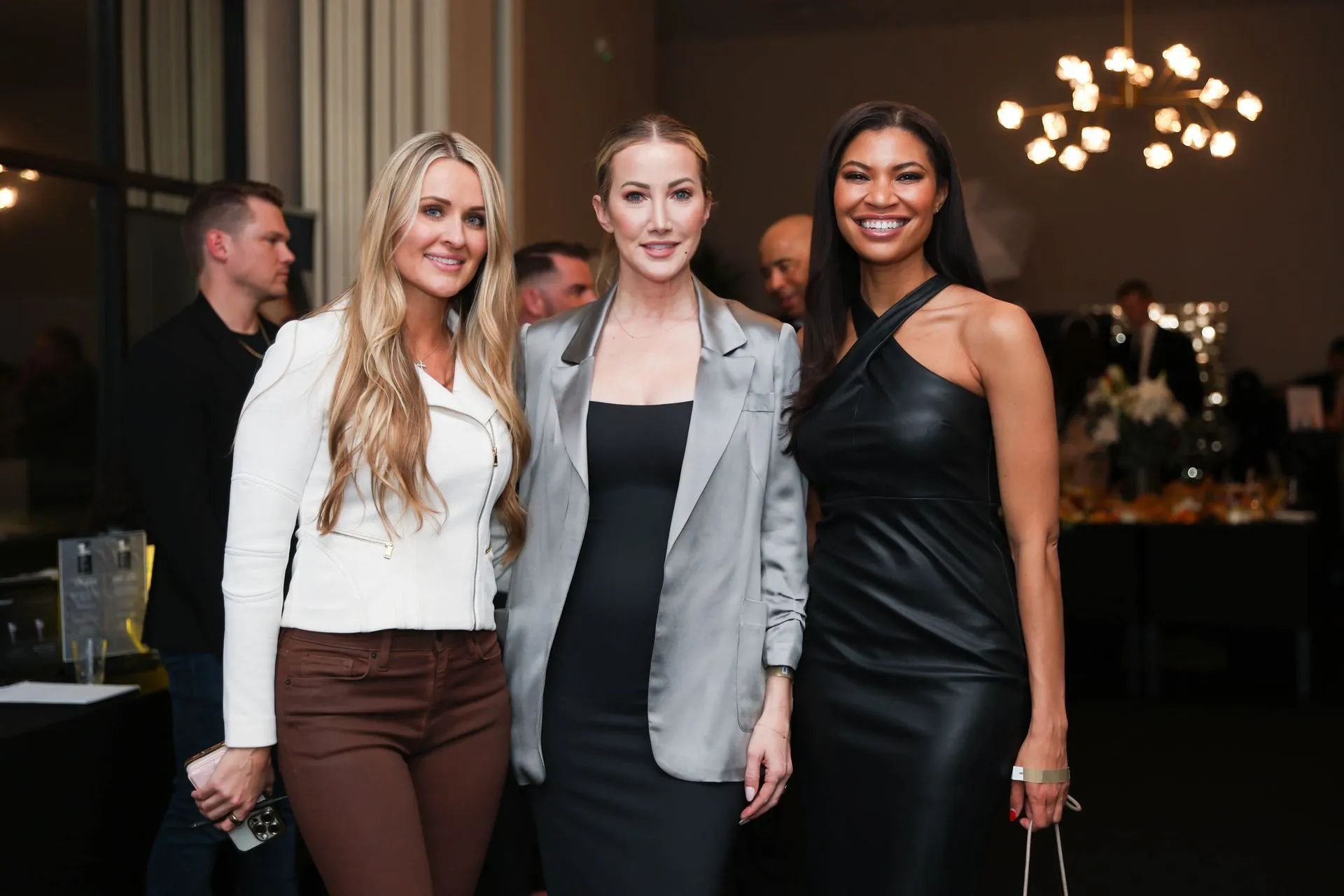 Three people posing indoors at a formal event, two in dresses and one in a blazer, with chandeliers behind them.