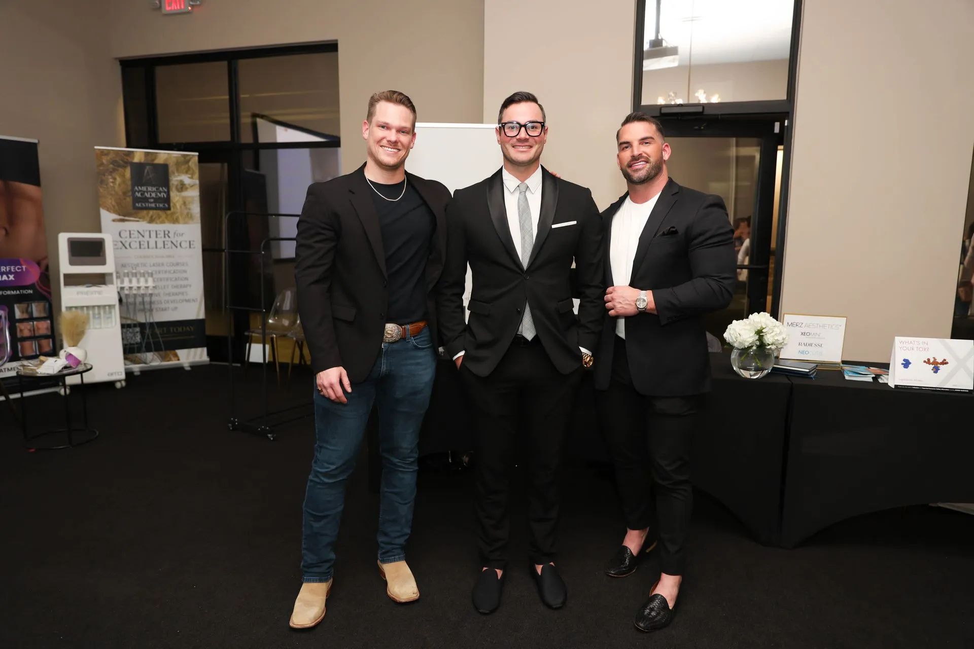 Three men in black formalwear pose at an indoor event table with brochures and a flower arrangement.