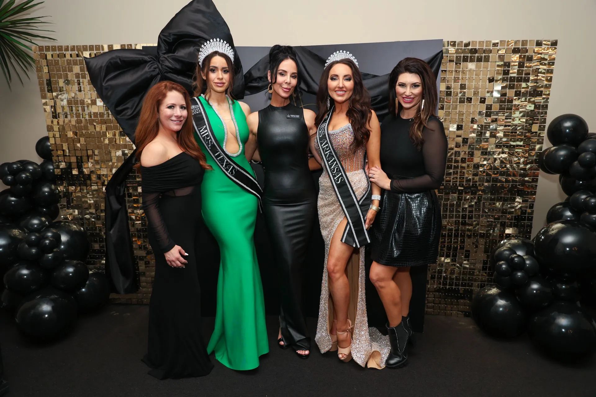 Five women posing at a party in formal dresses with black balloon decorations and a gold backdrop.