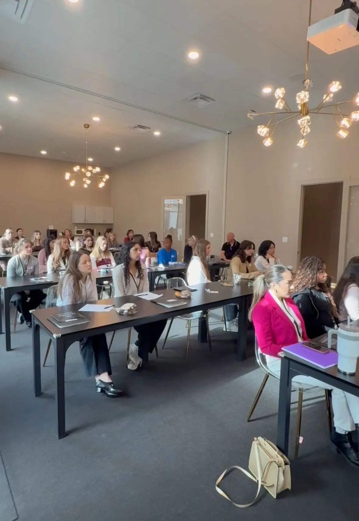 Conference room with attendees seated at tables, listening during a meeting under hanging chandeliers.