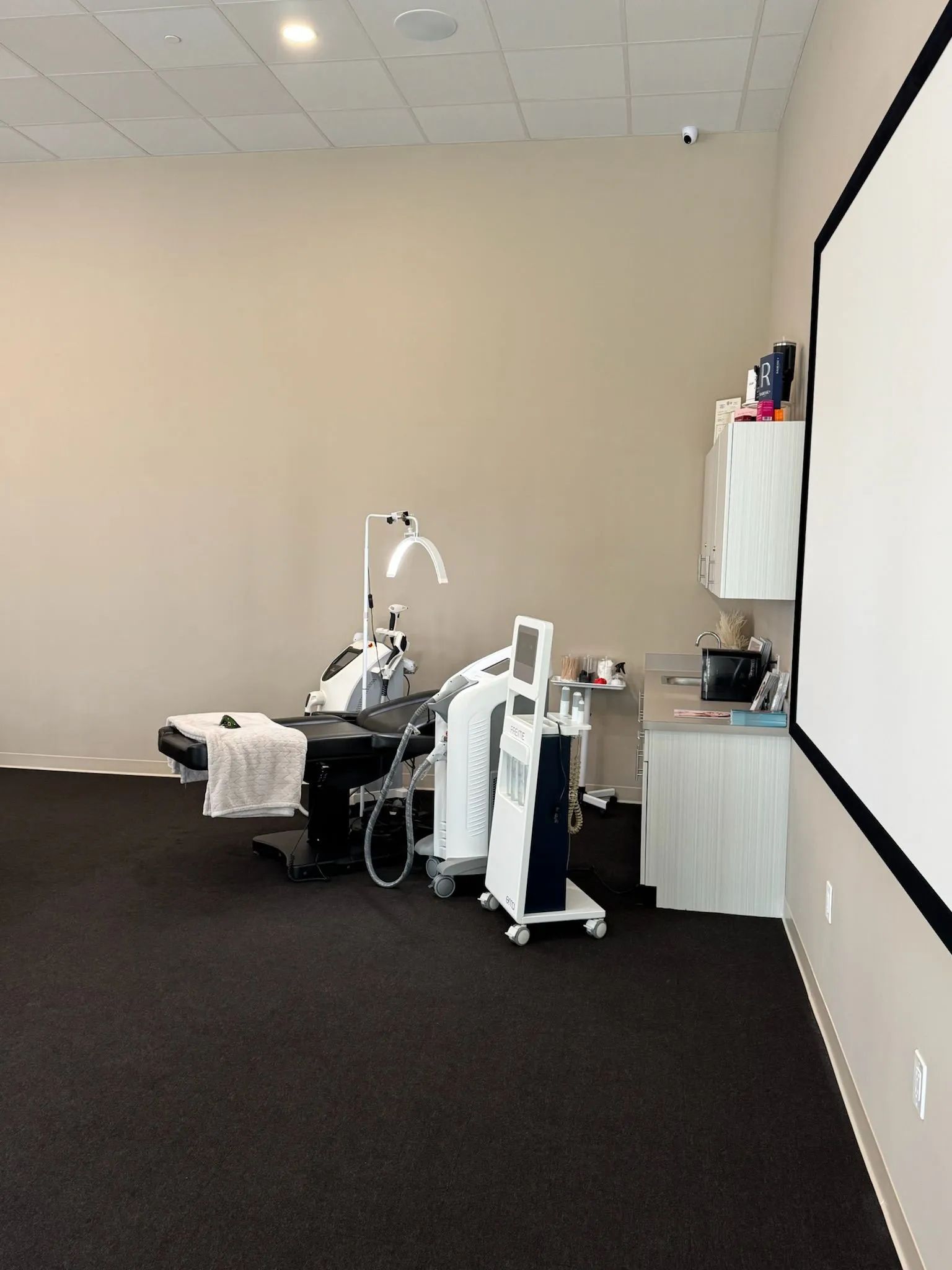 Empty clinical room with medical equipment carts, chair, and a wall-mounted cabinet.
