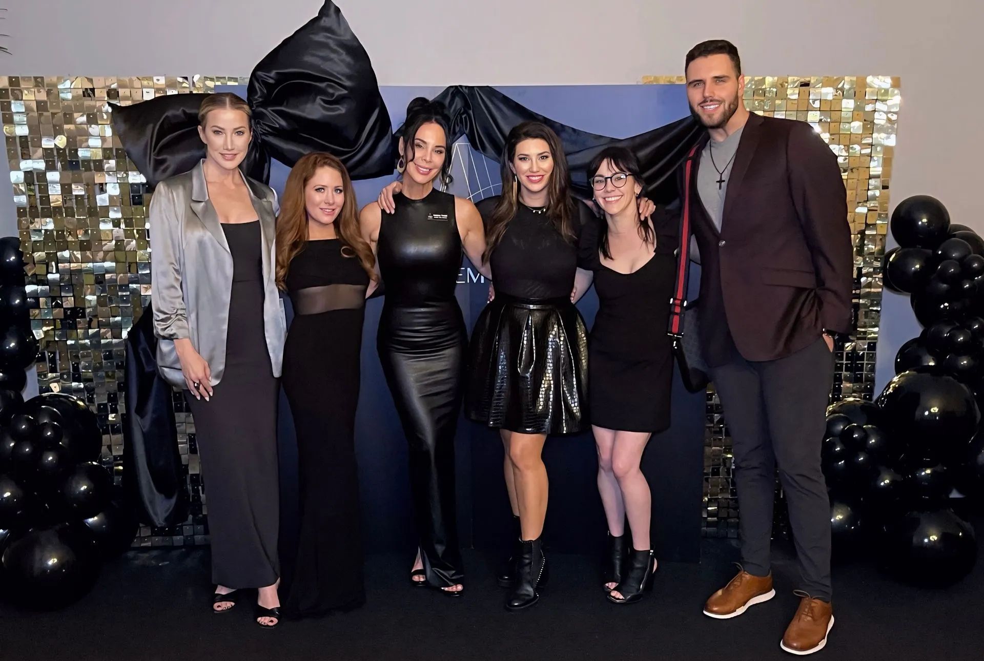 Six people posing at a black-and-gold party backdrop with balloons and a large black bow