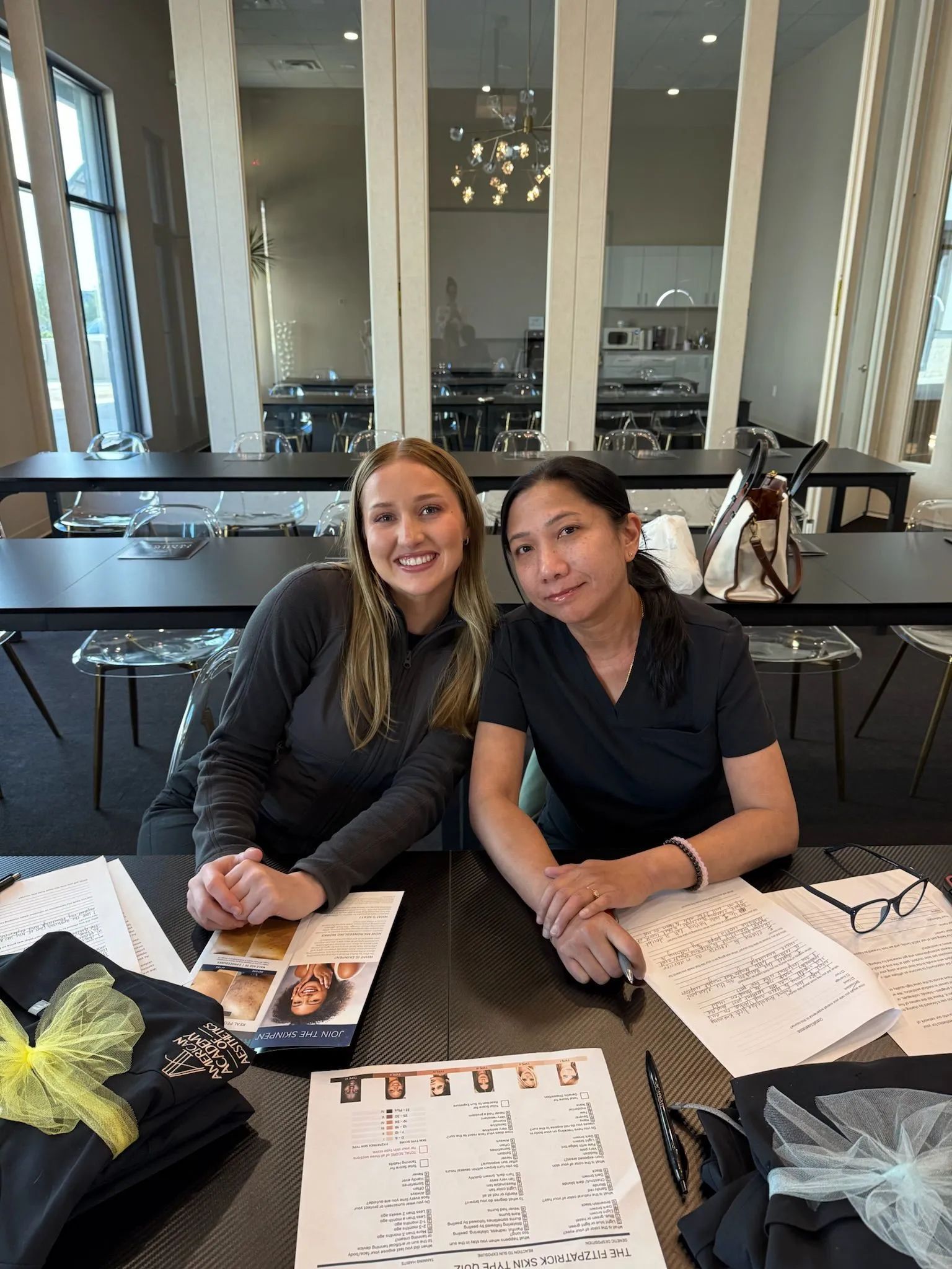 Two people smiling at a table with papers and a tote bag in a bright room with mirrored walls