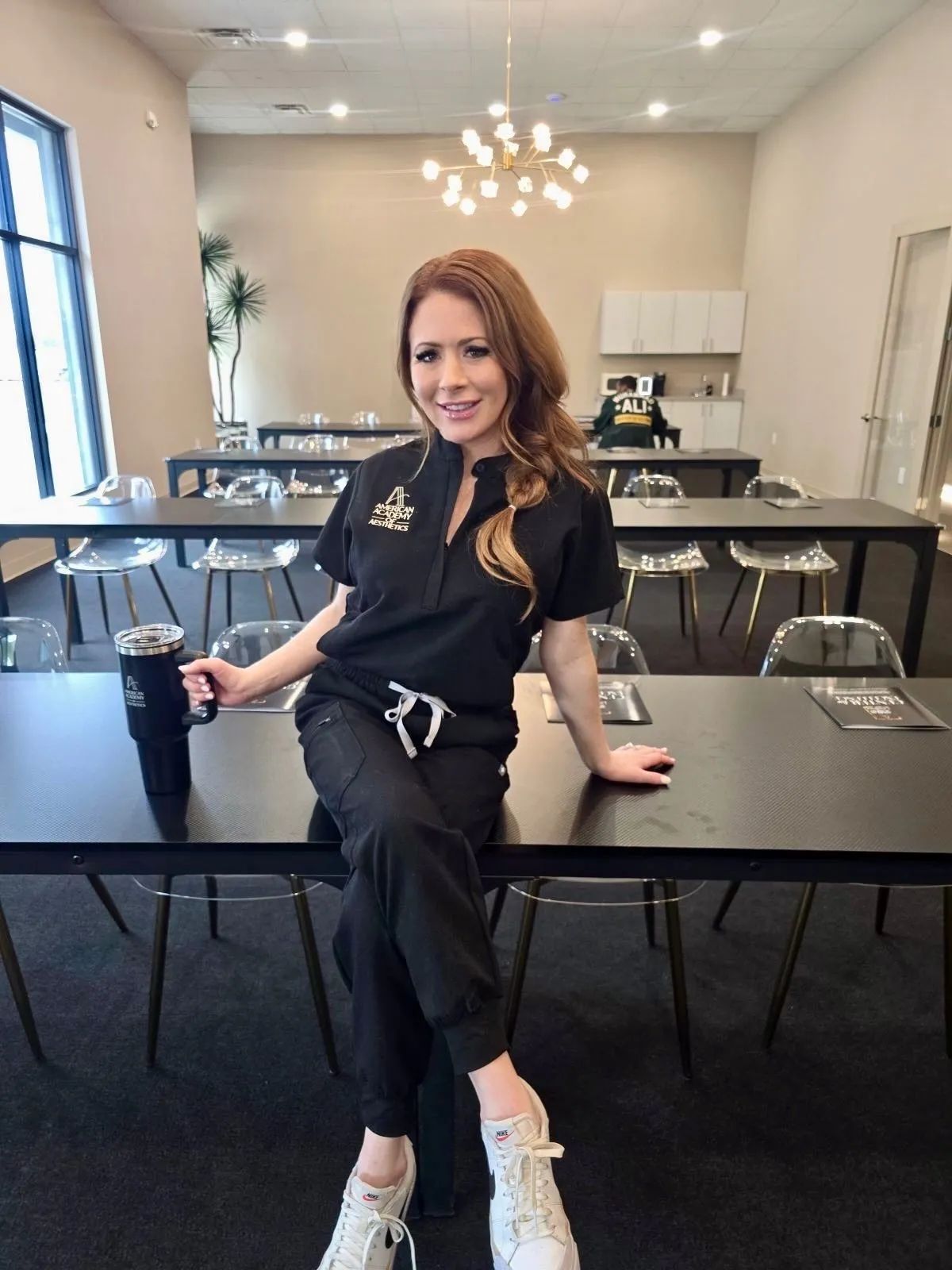 Woman in black outfit sitting on a conference table in a bright meeting room, smiling at the camera.