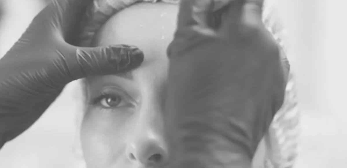 Gloved hands shaping a woman’s eyebrows during a makeup or beauty treatment