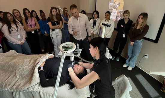 Group gathered around a bed as a woman receives makeup or skincare treatment indoors.