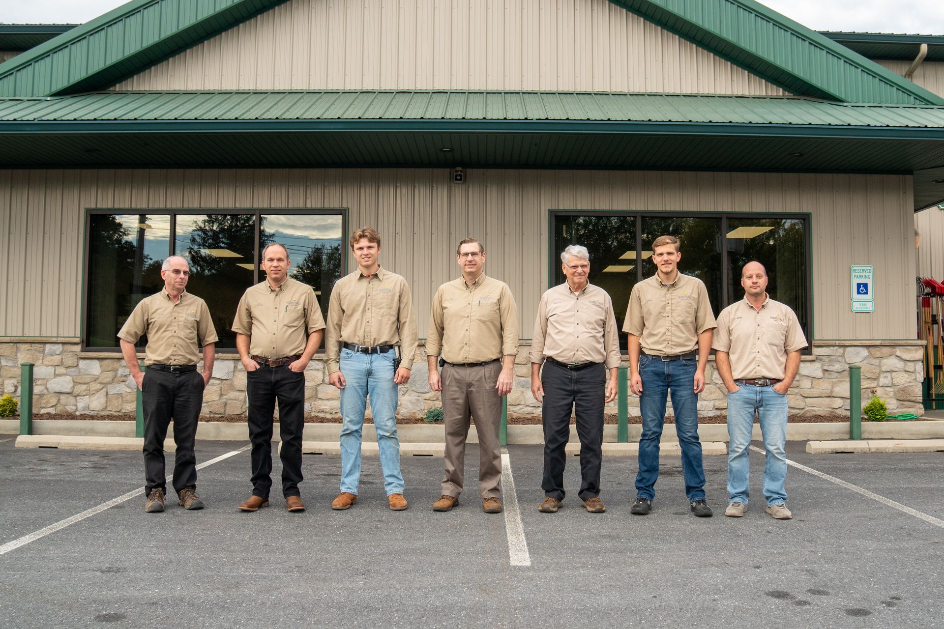 Seven people stand in a line in front of a building. They wear similar tan shirts and various dark pants and jeans.