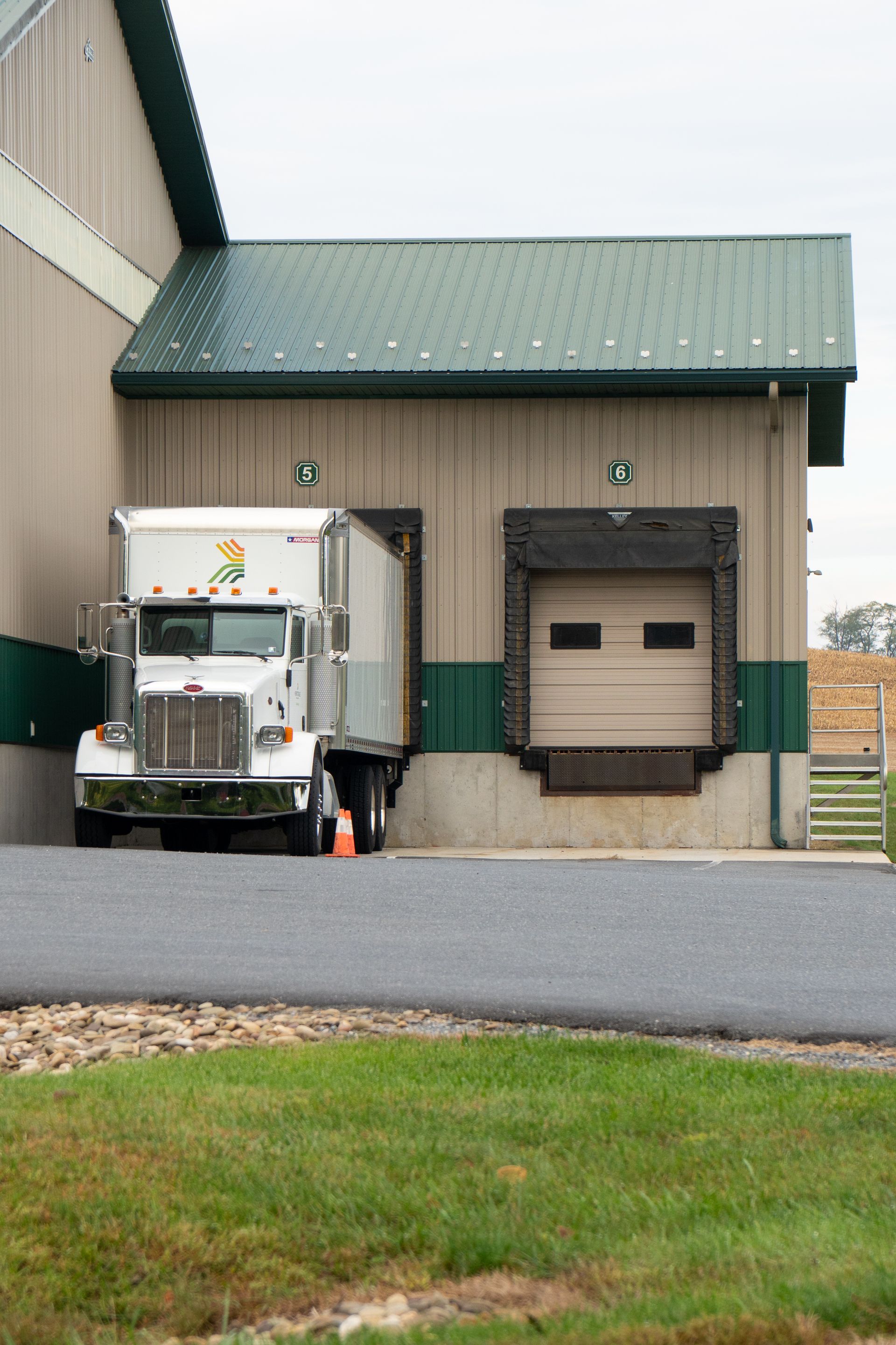White semi-truck at a loading dock with closed door. Building has green trim and roof.
