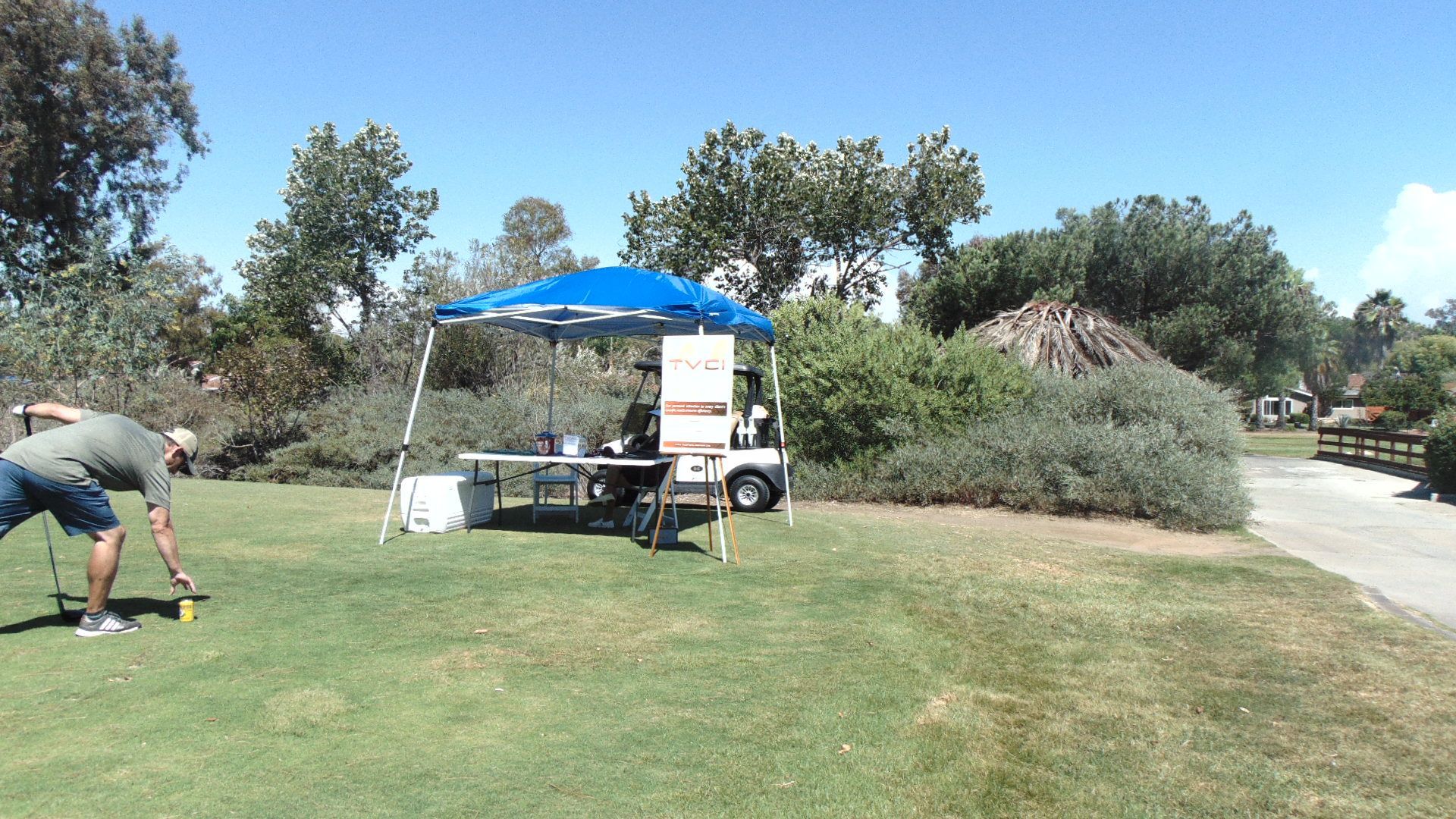 Person setting up a tent beside a blue canopy in a sunny park near a road