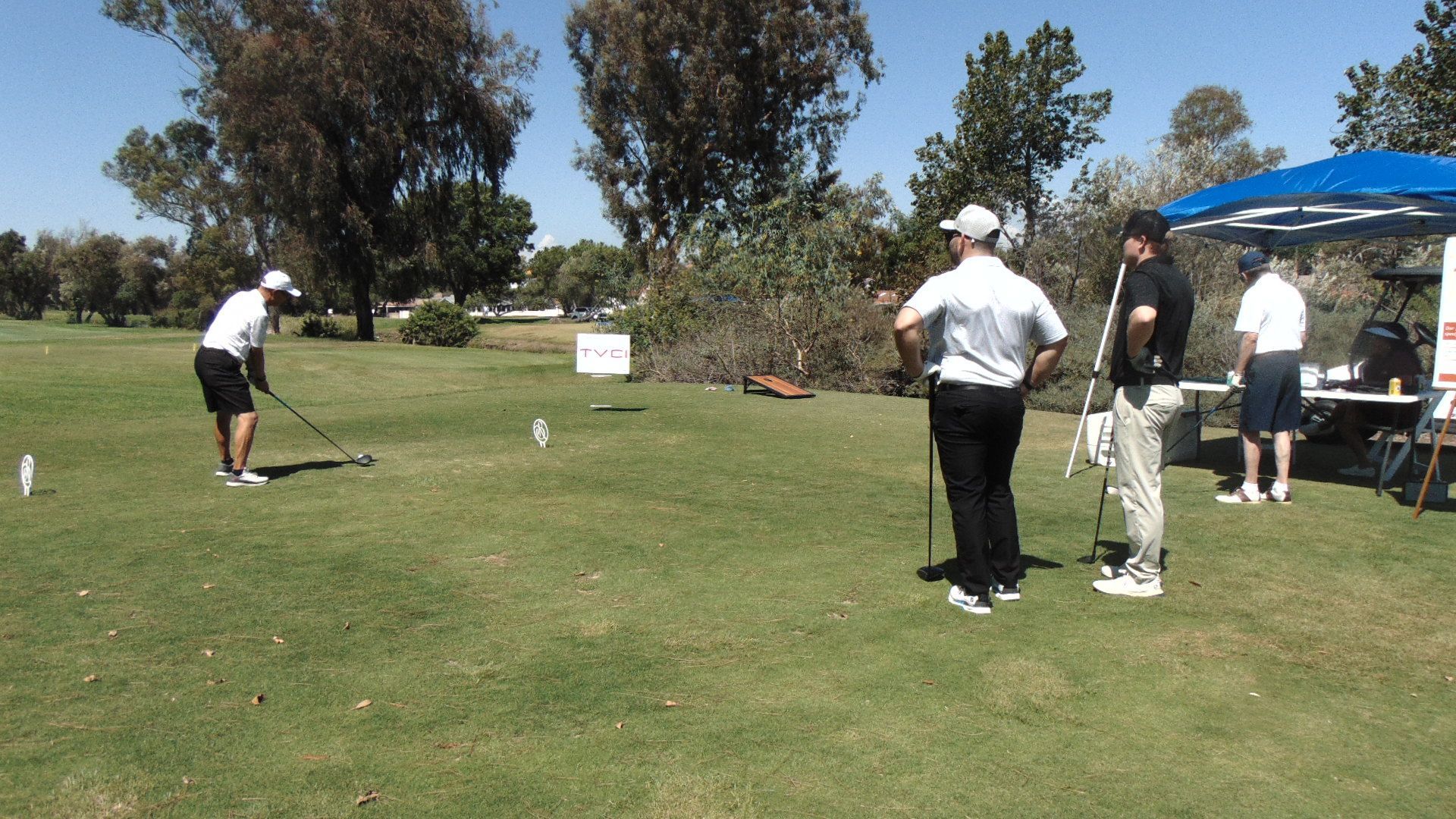 People playing golf on a sunny grassy course near a blue canopy tent.