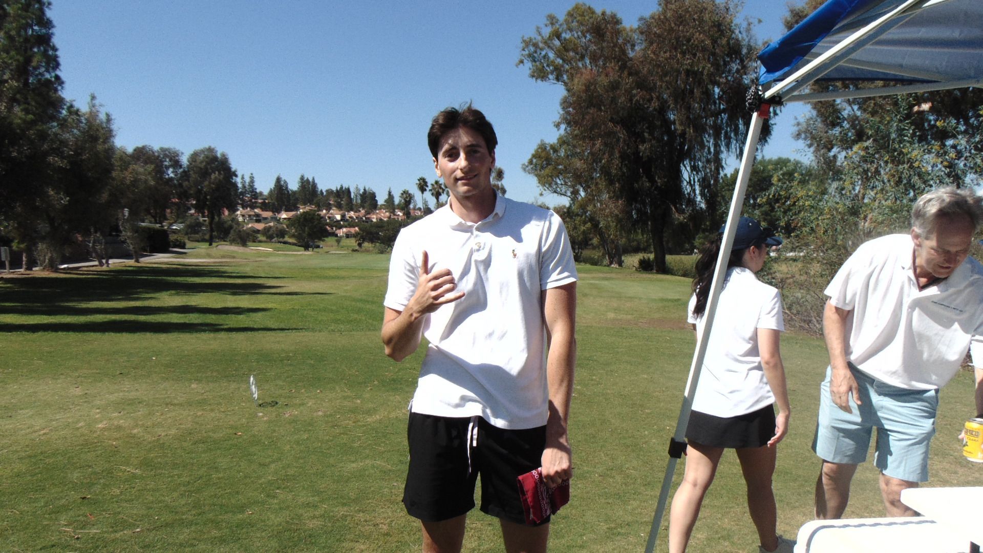 People standing on a sunny golf course near a blue canopy, with one person holding a drink.