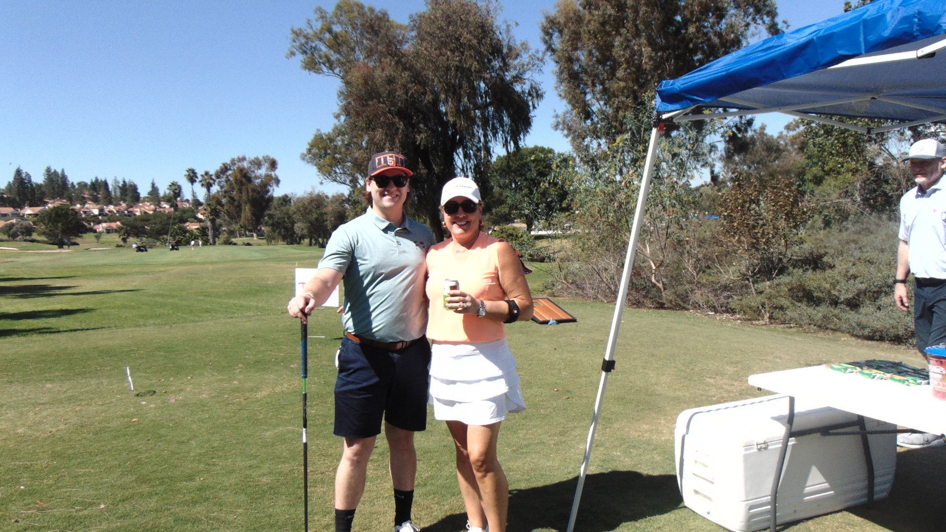 Two golfers posing together on a sunny golf course beside a blue canopy tent.
