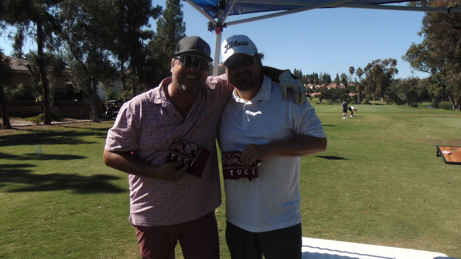 Two people pose together under a shade structure on a sunny golf course.