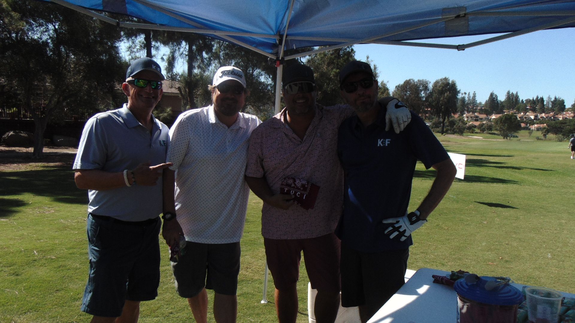 Four people standing together on a sunny golf course, with trees and fairways in the background.