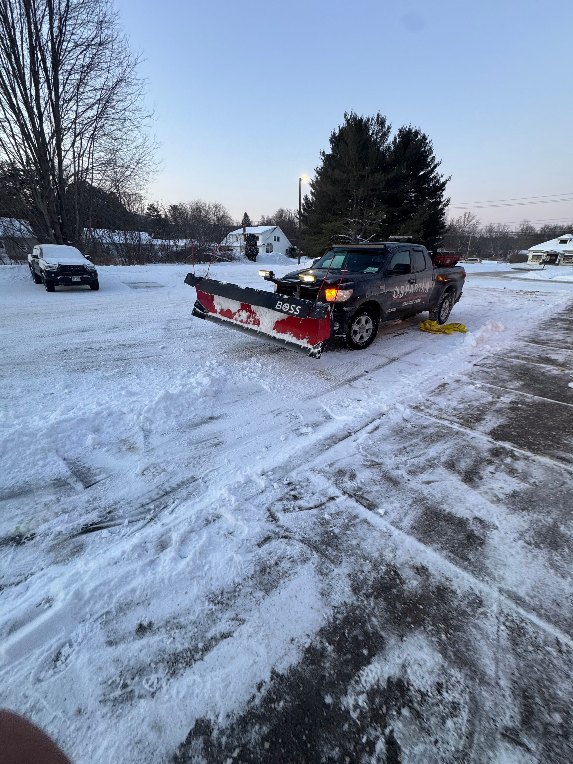 White pickup truck with snowplow, parked on gravel, another truck in background. 
