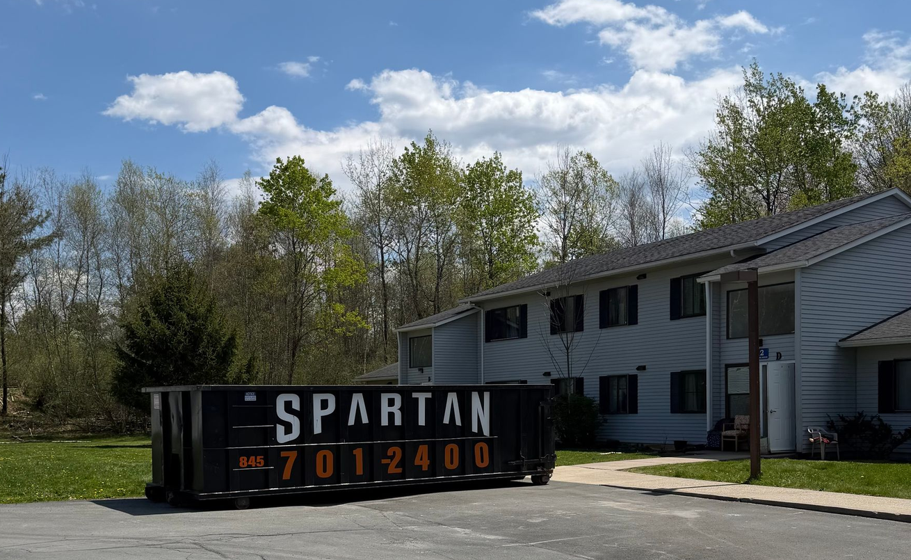 Black SPARTAN dumpster in a parking lot beside a white building under a blue sky.