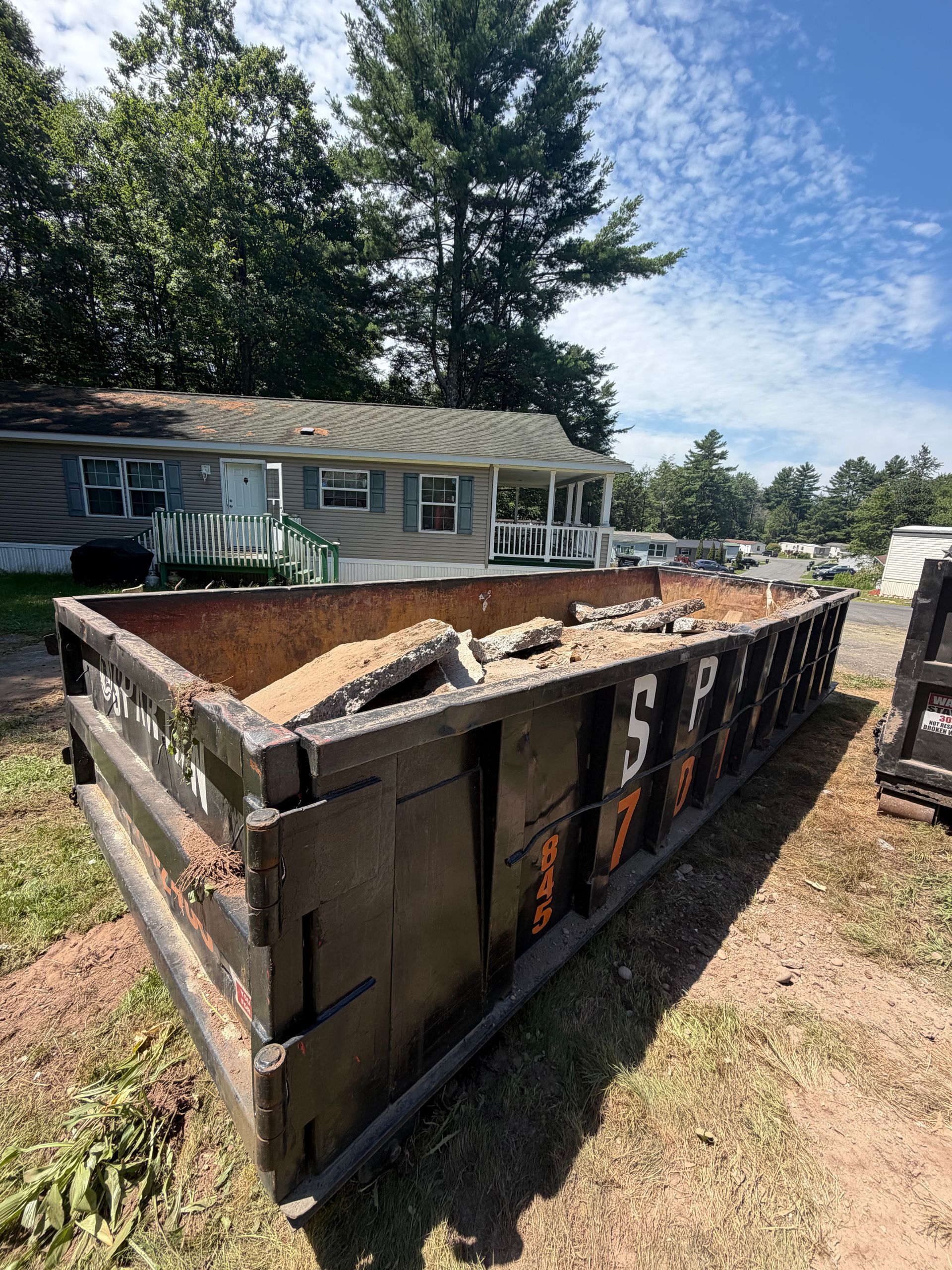 Large metal dumpster filled with debris beside a white house and trees on a sunny day