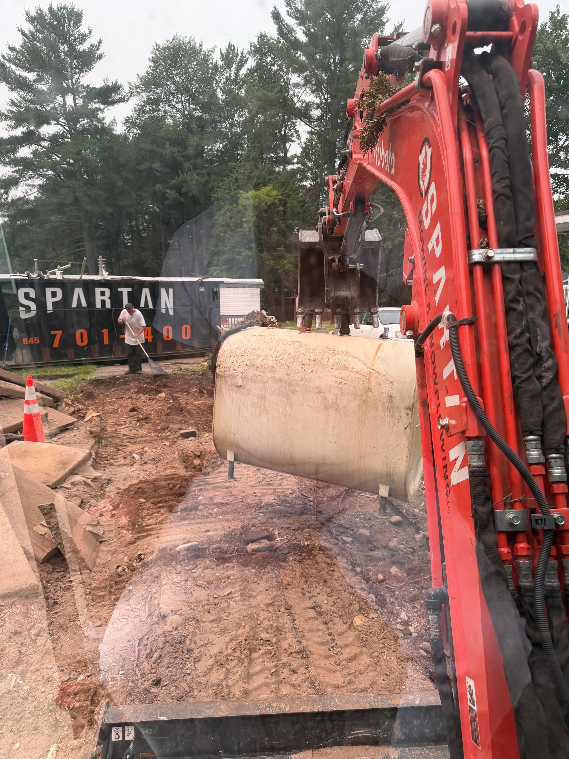 Red excavator lifting a large stone block at a construction site with dirt piles and trees nearby.