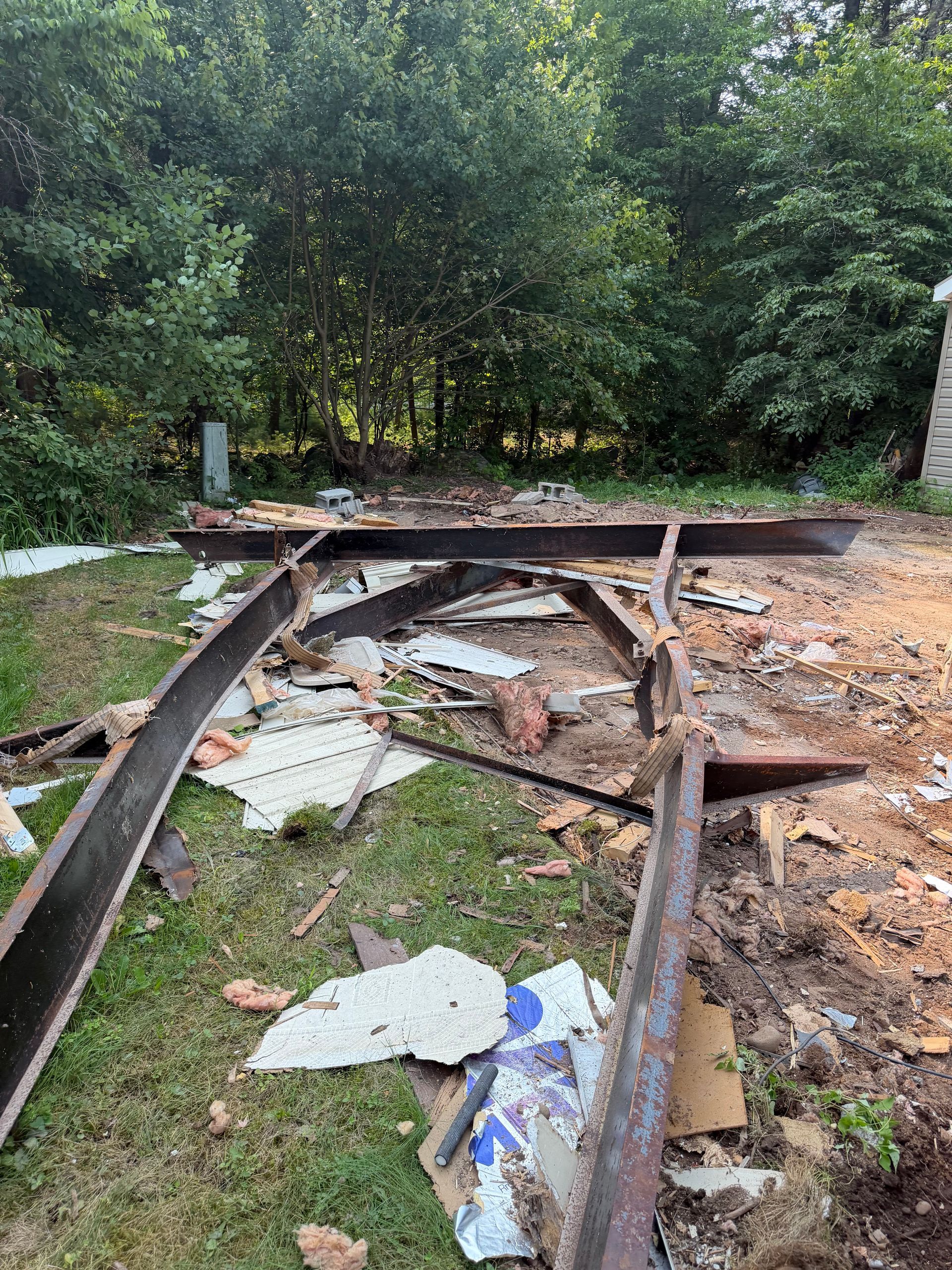 Collapsed wooden frame with scattered debris on a muddy ground beside dense green trees