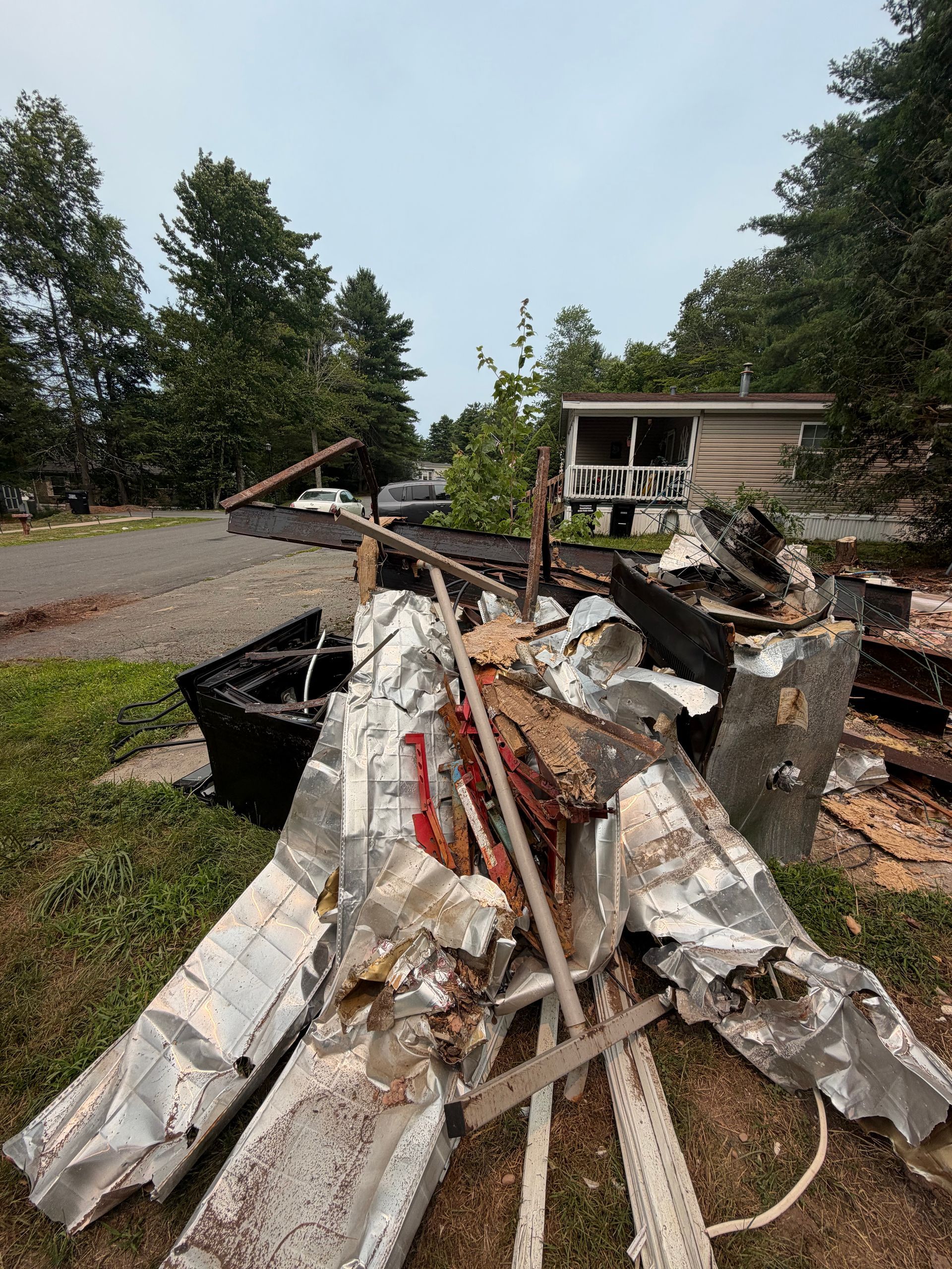 Burned debris and twisted metal piled by a roadside near a house and trees
