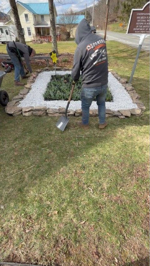Two people landscaping a rectangular garden bed with white rocks and greenery near a historic marker and a house.
