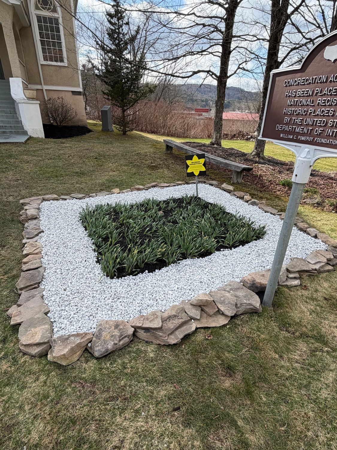 A rectangular memorial garden with white stone borders and a central planting bed, next to a historical marker sign.